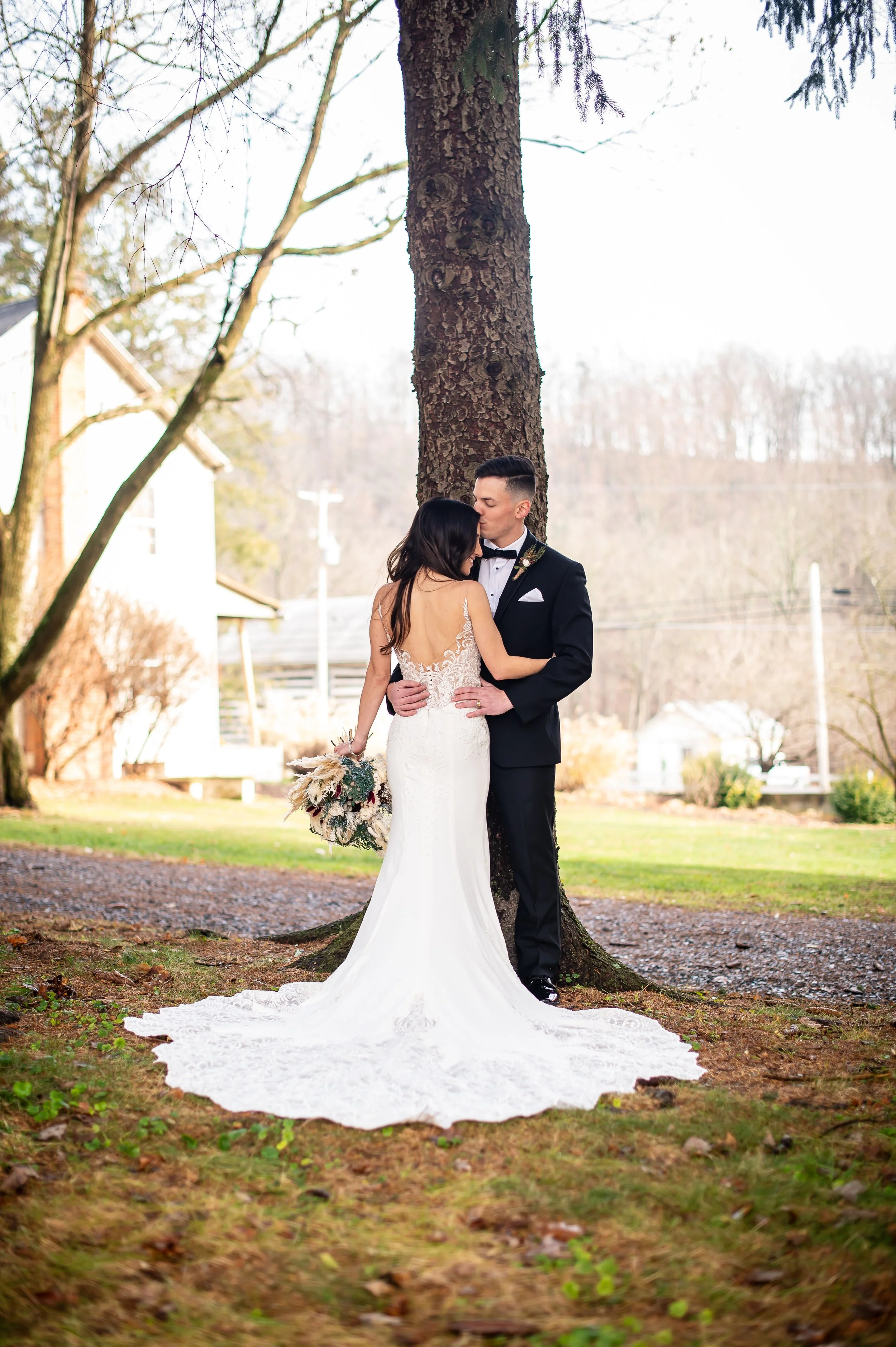 A bride and groom embrace beneath a large tree outdoors, with the groom kissing the bride's forehead. The bride wears a white lace wedding gown with a train, and the groom wears a black tuxedo with a bowtie. The bride holds a bouquet of dried flowers