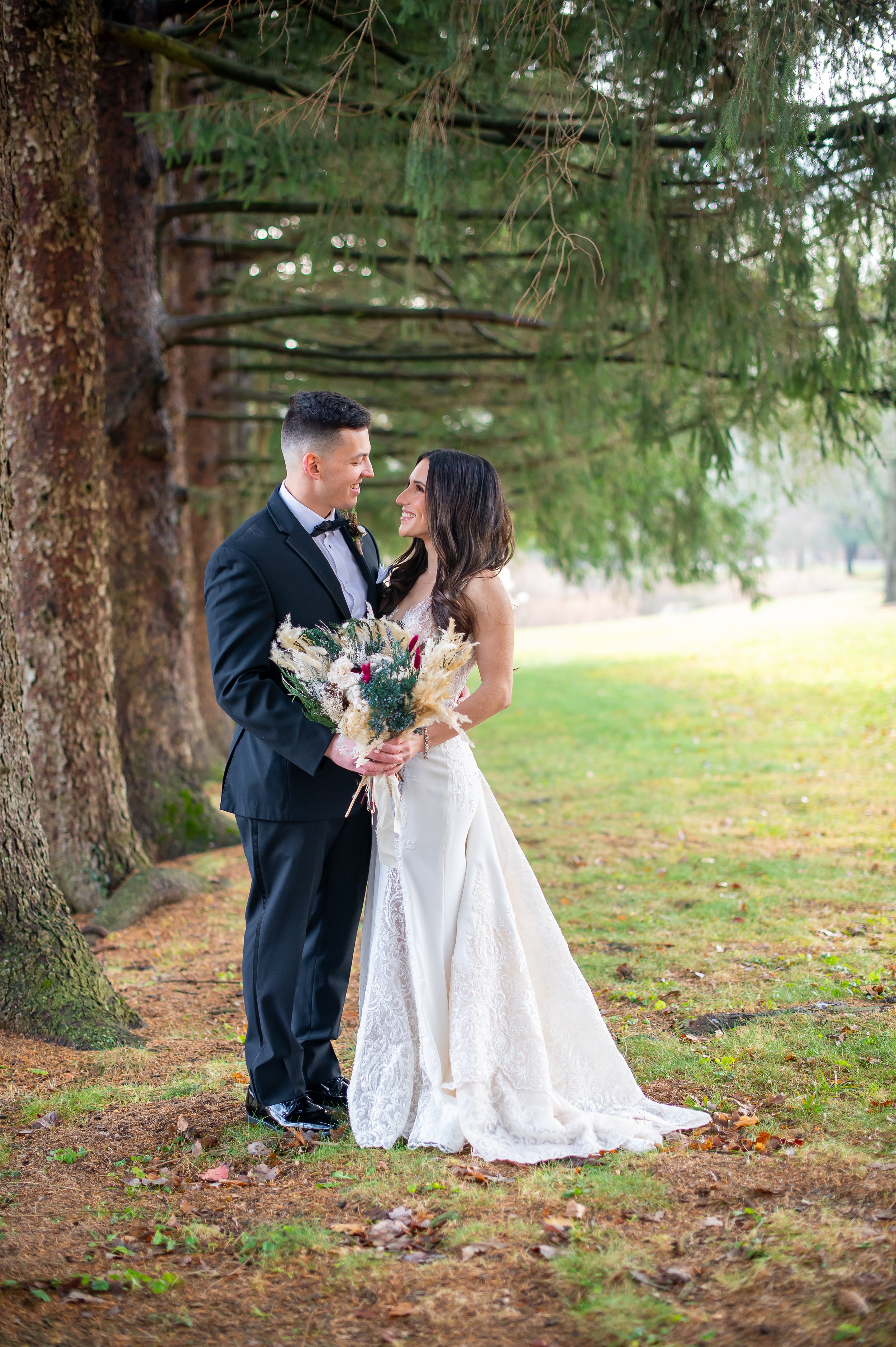 A newlywed couple stands together outdoors in a forested area on their wedding day, smiling at each other. The groom wears a black tuxedo with a bow tie, and the bride wears a white lace wedding dress holding a bouquet of dried flowers and greenery.