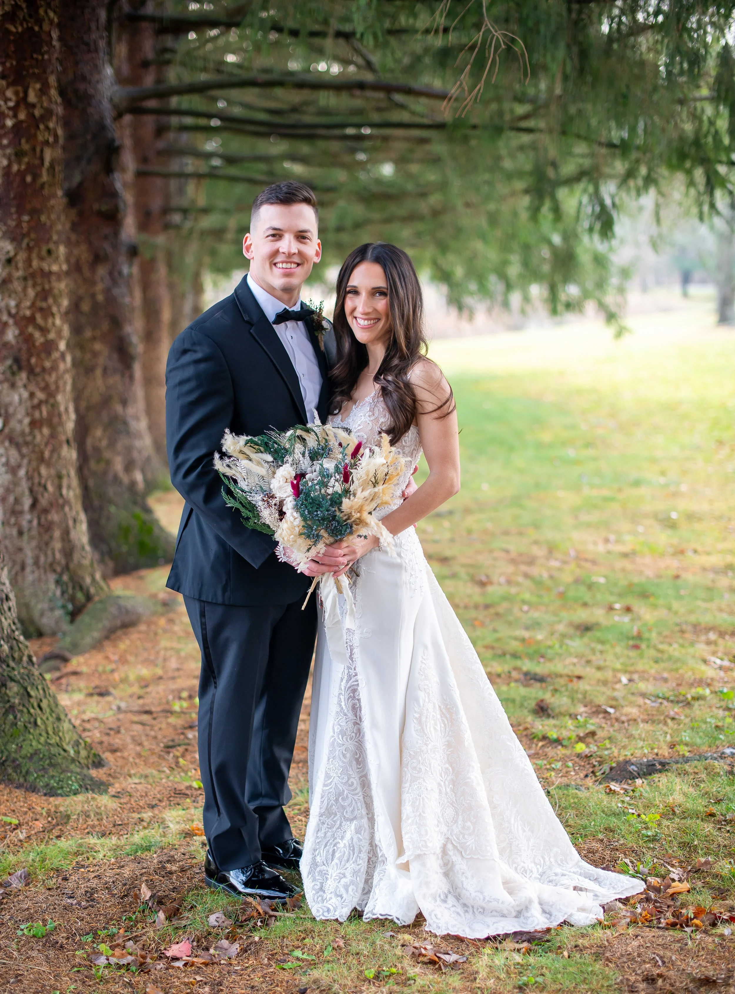 A newlywed couple standing together outdoors near a tree, smiling, with the groom in a black tuxedo and the bride in a white lace wedding gown holding a bouquet of flowers.
