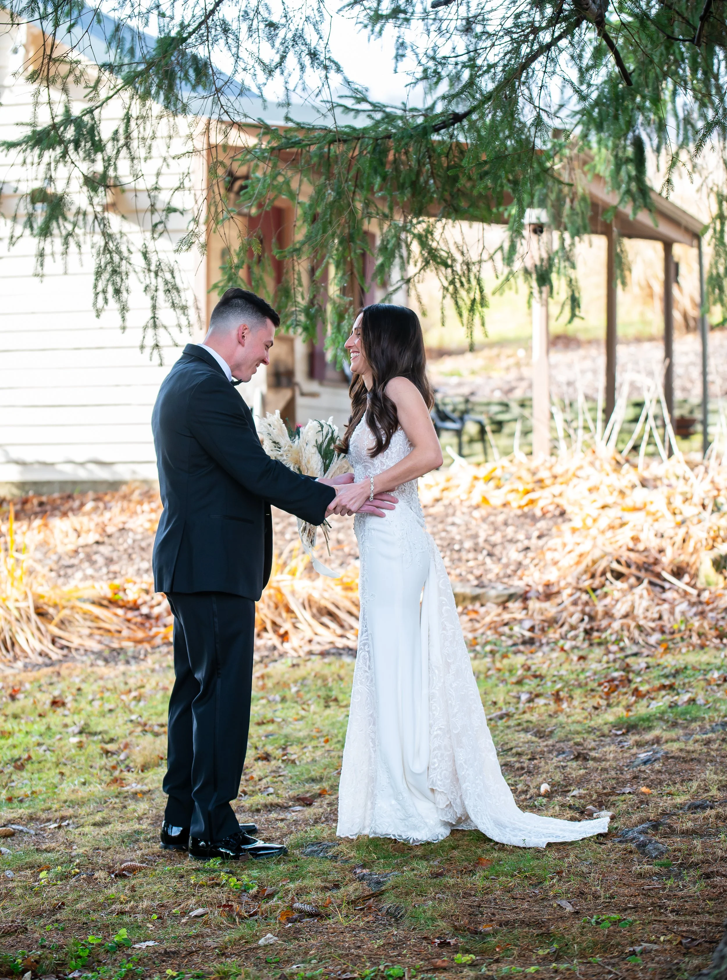 A bride and groom stand outside under a tree, holding hands and smiling during their wedding ceremony, with a house and autumn leaves in the background.