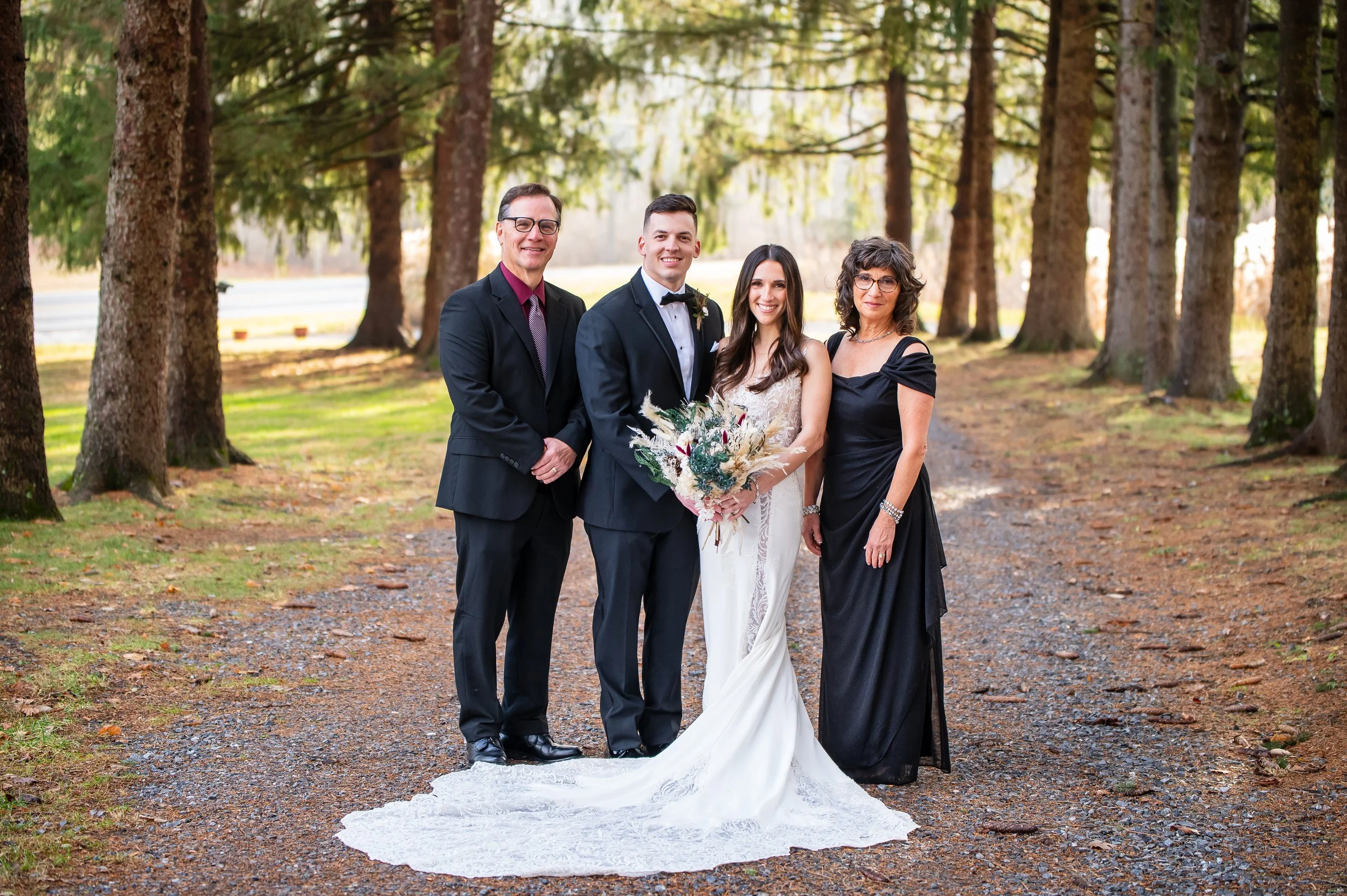 A wedding party standing outdoors on a dirt path surrounded by tall trees, featuring a bride in a lace wedding dress holding a bouquet, a groom in a tuxedo, and two guests in formal attire.