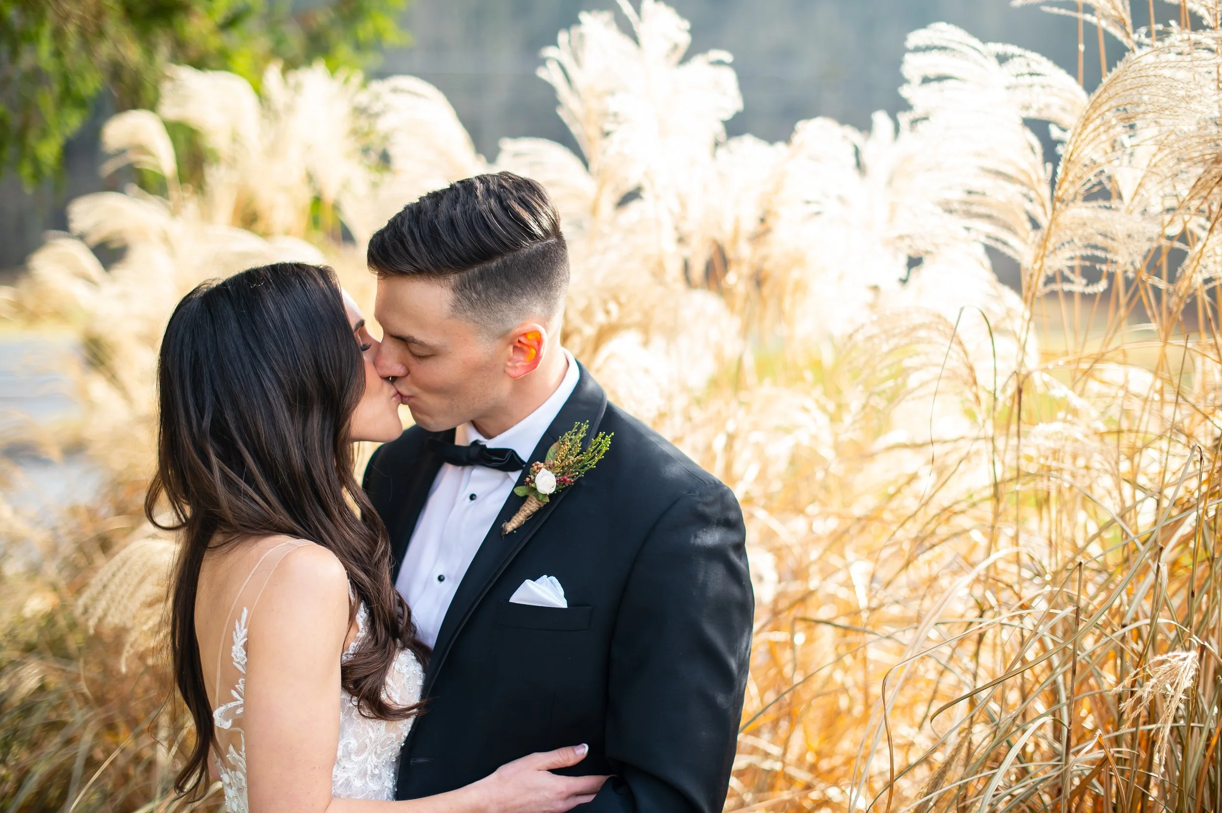 A bride and groom kiss outdoors in front of tall dry grasses during a wedding ceremony.