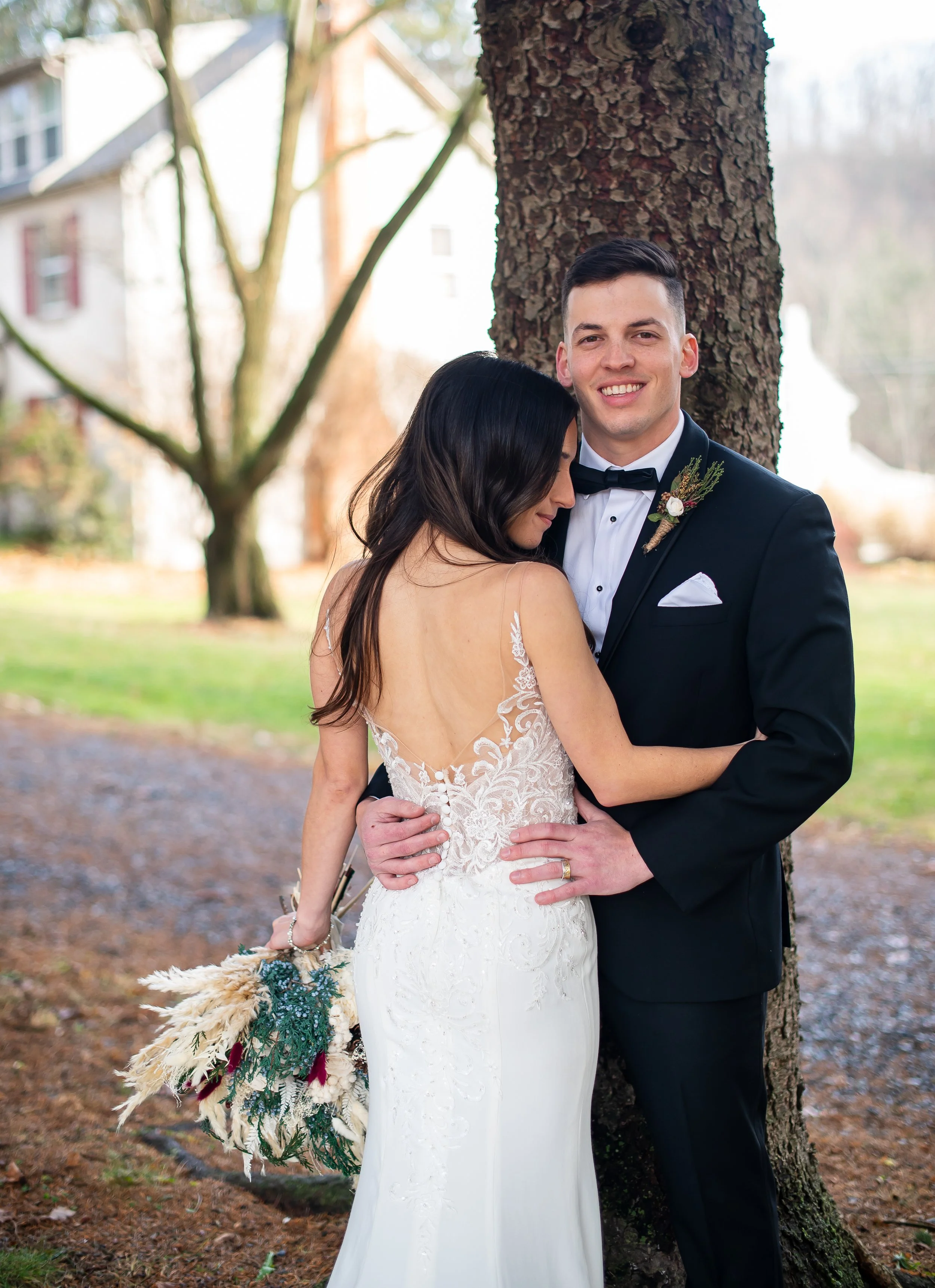 A bride and groom embrace outdoors during daytime. The bride has dark hair and wears a white wedding gown with lace back; the groom has short dark hair and is dressed in a black tuxedo with bow tie, standing next to a large tree.
