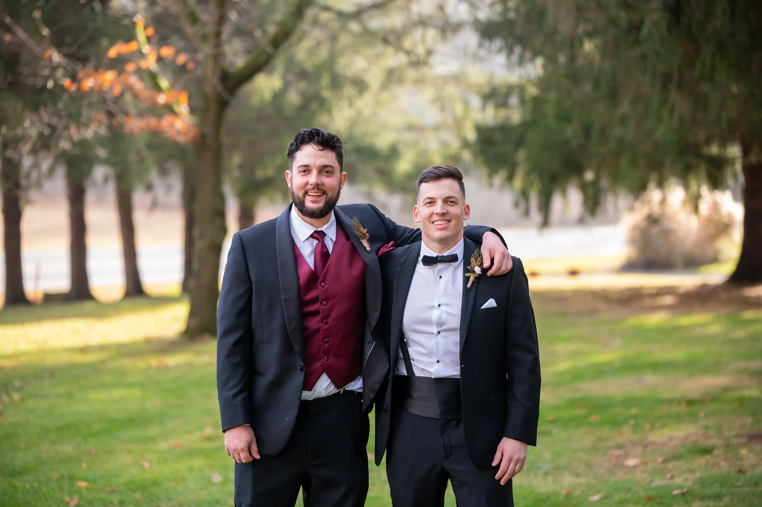 Two men in tuxedos smiling outdoors, with one man with his arm around the other, on a grassy area with trees and a body of water in the background.