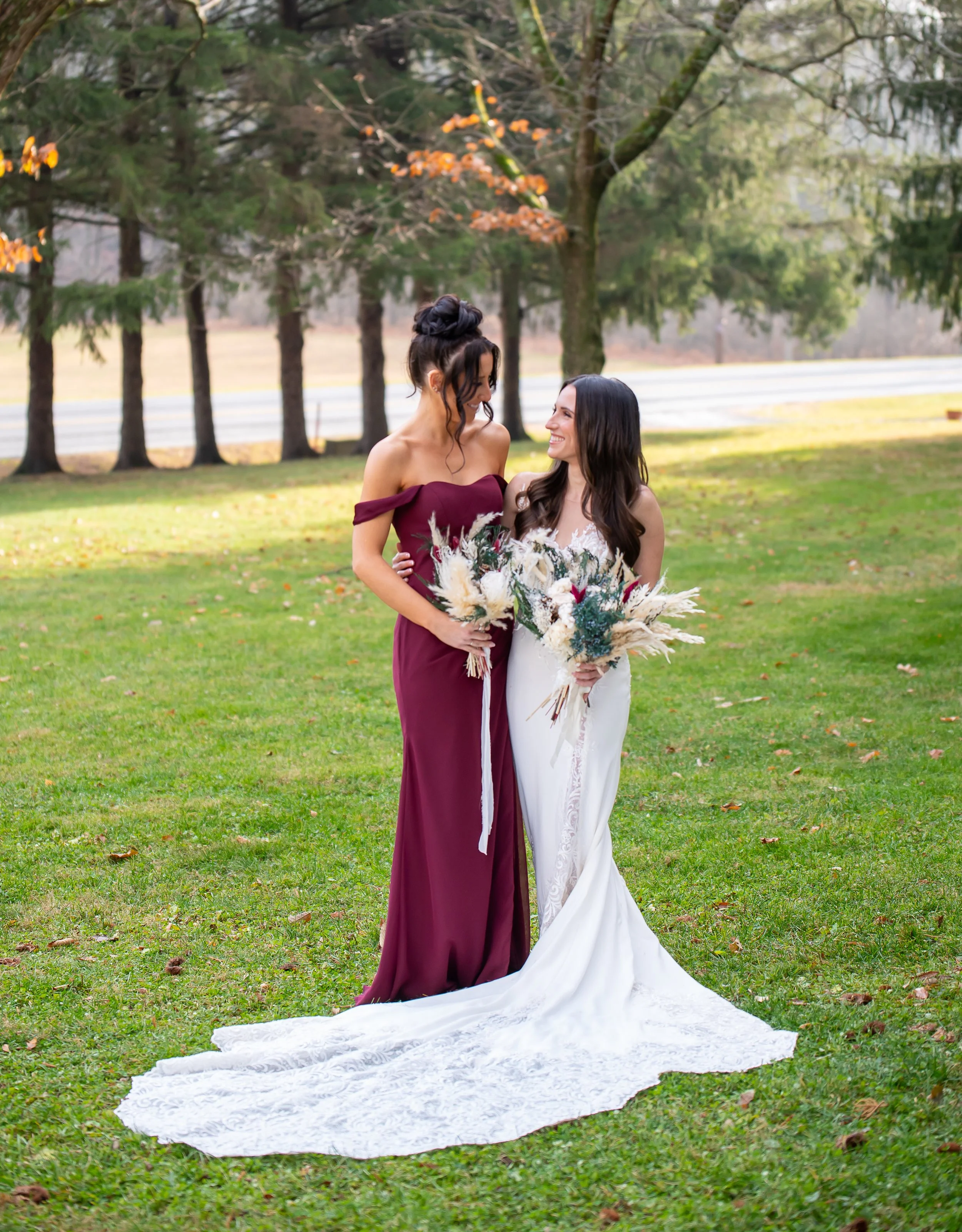 Two women in wedding attire standing on a grassy field, smiling at each other, holding bouquets, with trees in the background.