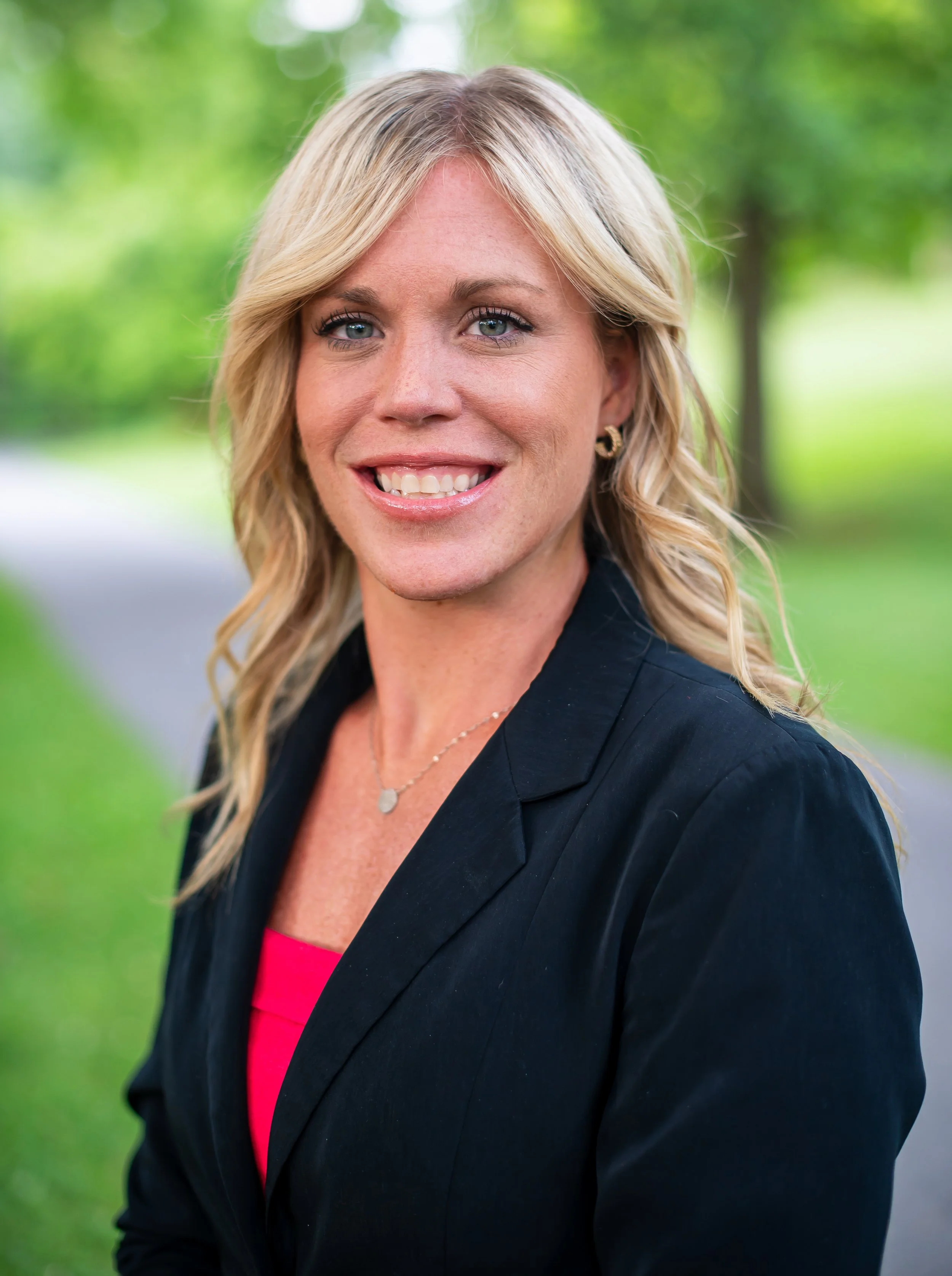 A woman with blonde hair and blue eyes smiling outdoors with a blurred green background, wearing a black blazer and red top.