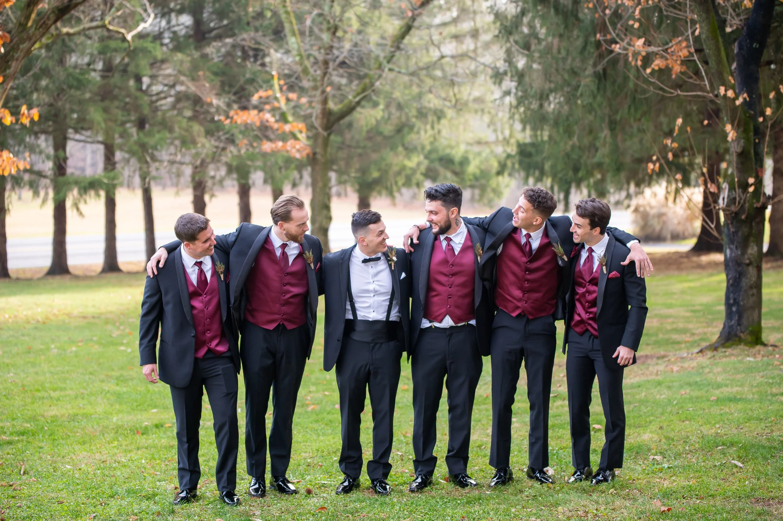 Group of seven men in tuxedos with maroon vests walking arm-in-arm outdoors in a park with trees and grass.