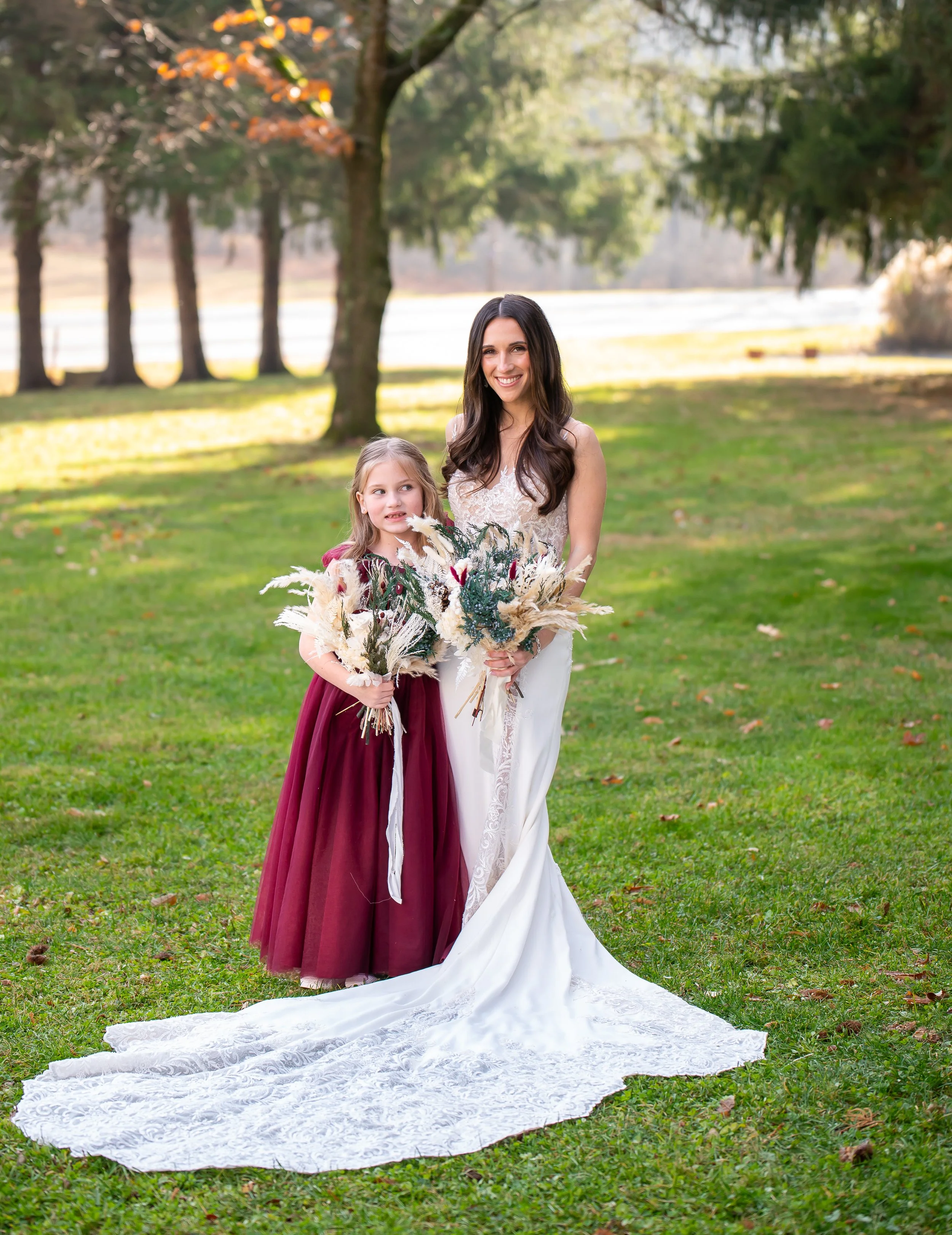 A woman in a white wedding dress holding a bouquet standing next to a young girl in a burgundy dress holding a bouquet, outdoors on a grassy field with trees in the background.