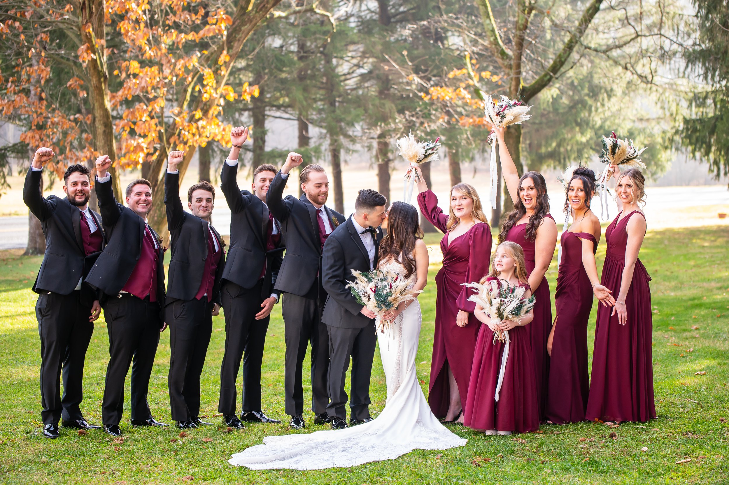 A group of wedding party members, including bride and groom, poses outdoors on a grassy field with trees in the background. The bridesmaids are in burgundy dresses and the groomsmen are in dark suits with burgundy vests. They are celebrating, some ra