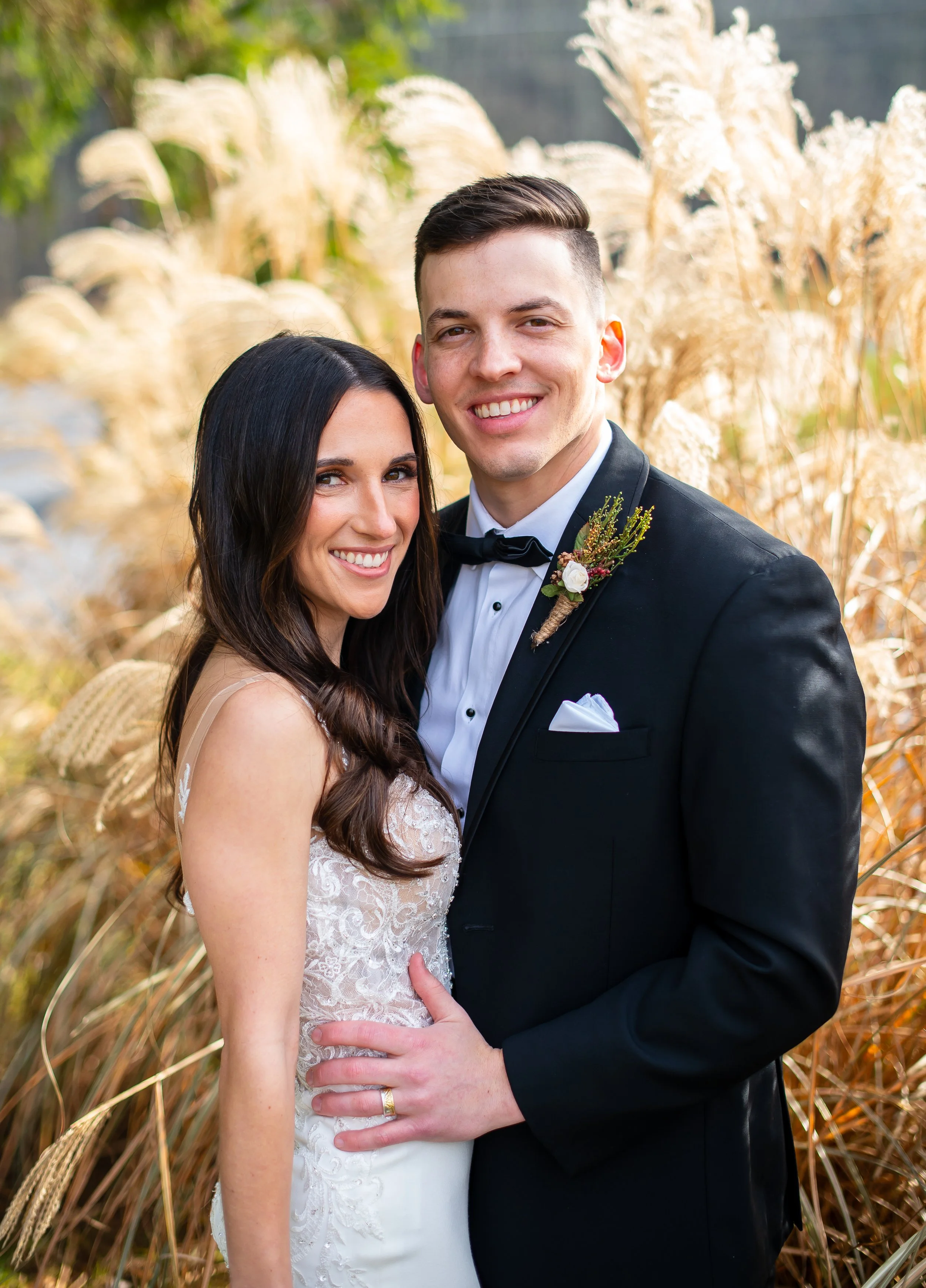 A newlywed couple standing outdoors among tall, golden grasses, smiling at the camera. The bride has long dark hair and is wearing a lace wedding dress, and the groom is in a black tuxedo with a bow tie, boutonniere, and white shirt.