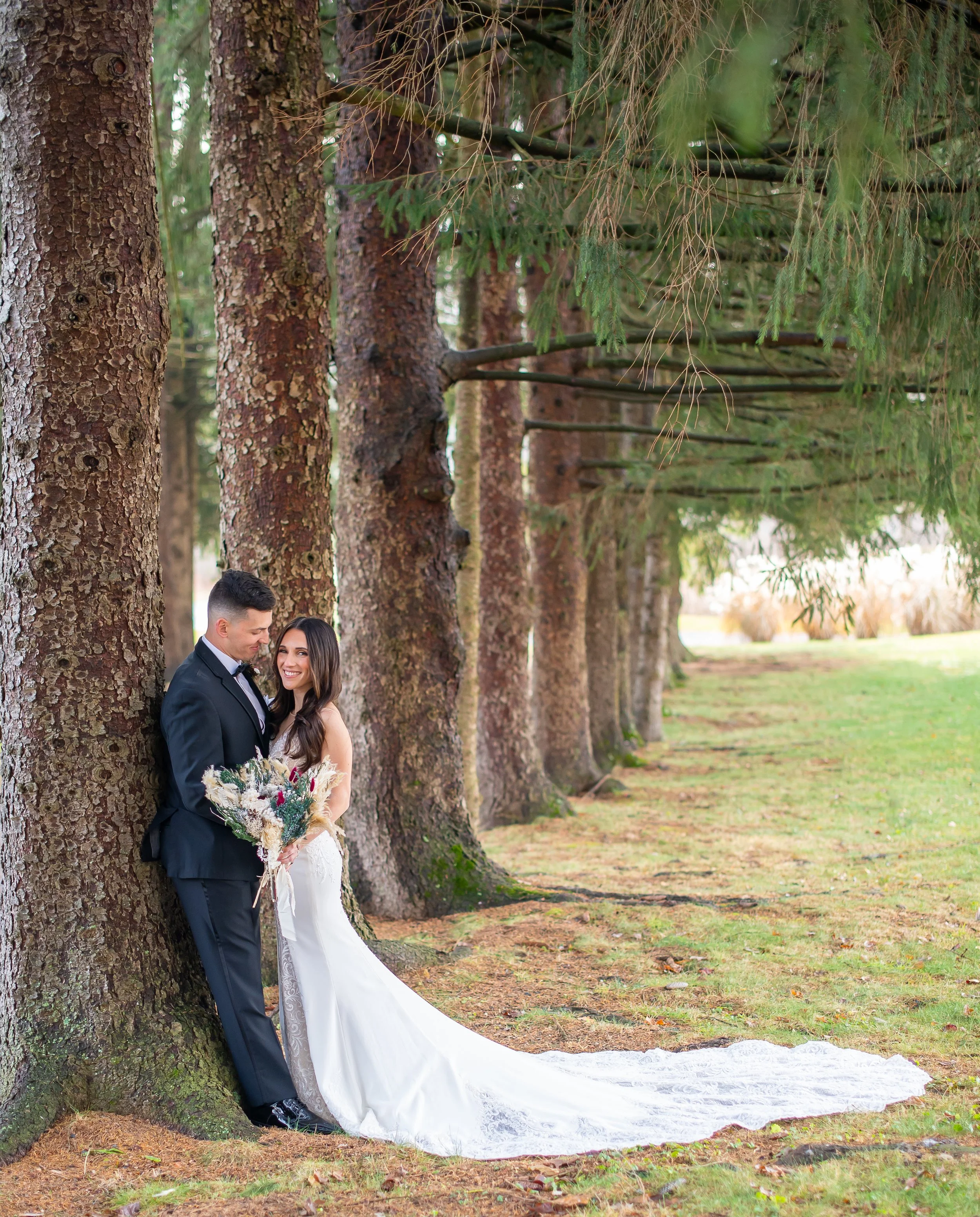A bride and groom standing close together next to large trees in a wooded area. The bride wears a white wedding dress with a long train and holds a bouquet of flowers; the groom is in a black suit and tie. They are smiling at each other, sharing an i