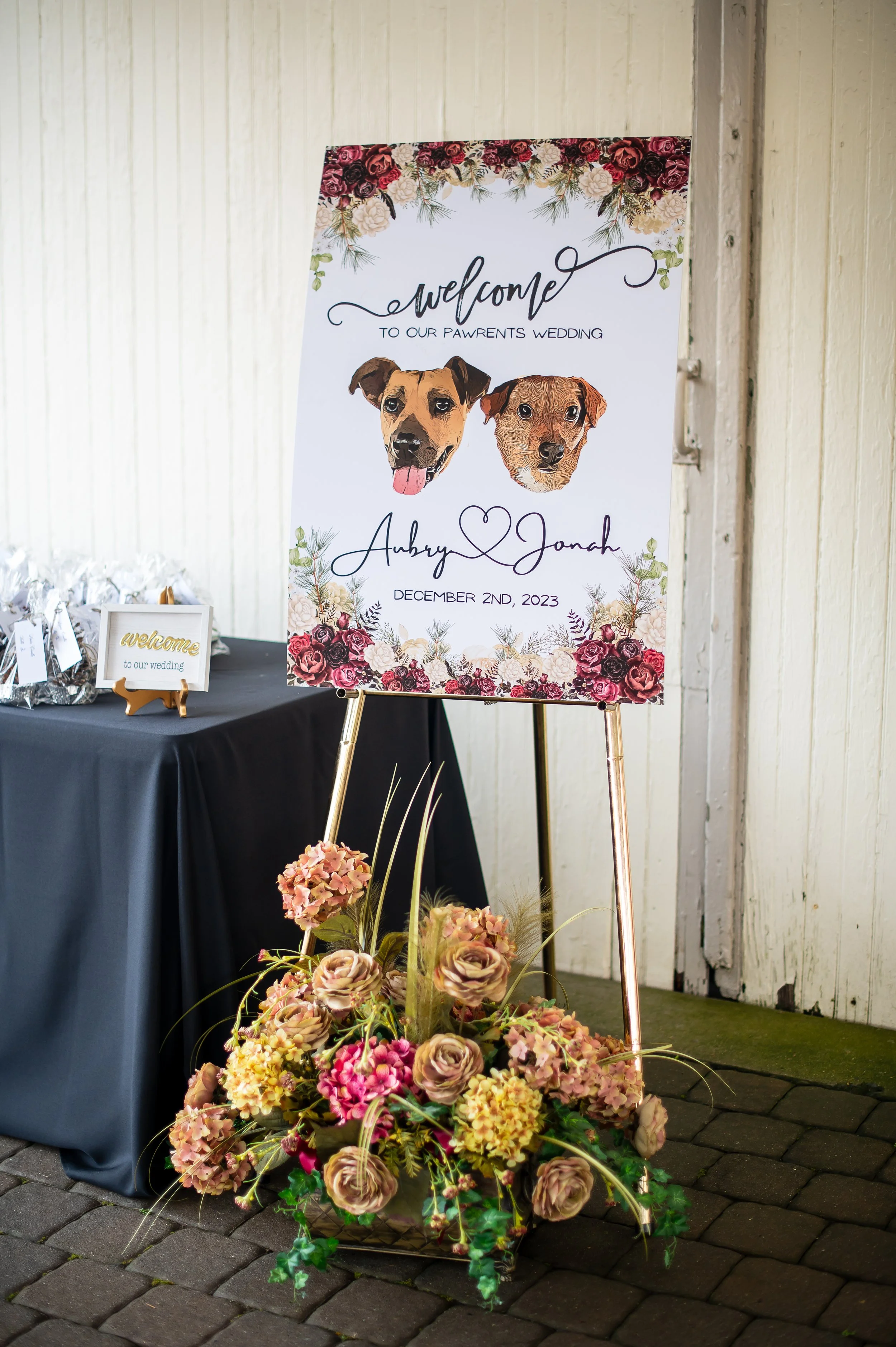 Wedding welcome sign with dog illustrations, floral decorations, and floral arrangement at the base, set against a white wall and black table with small welcome signage.