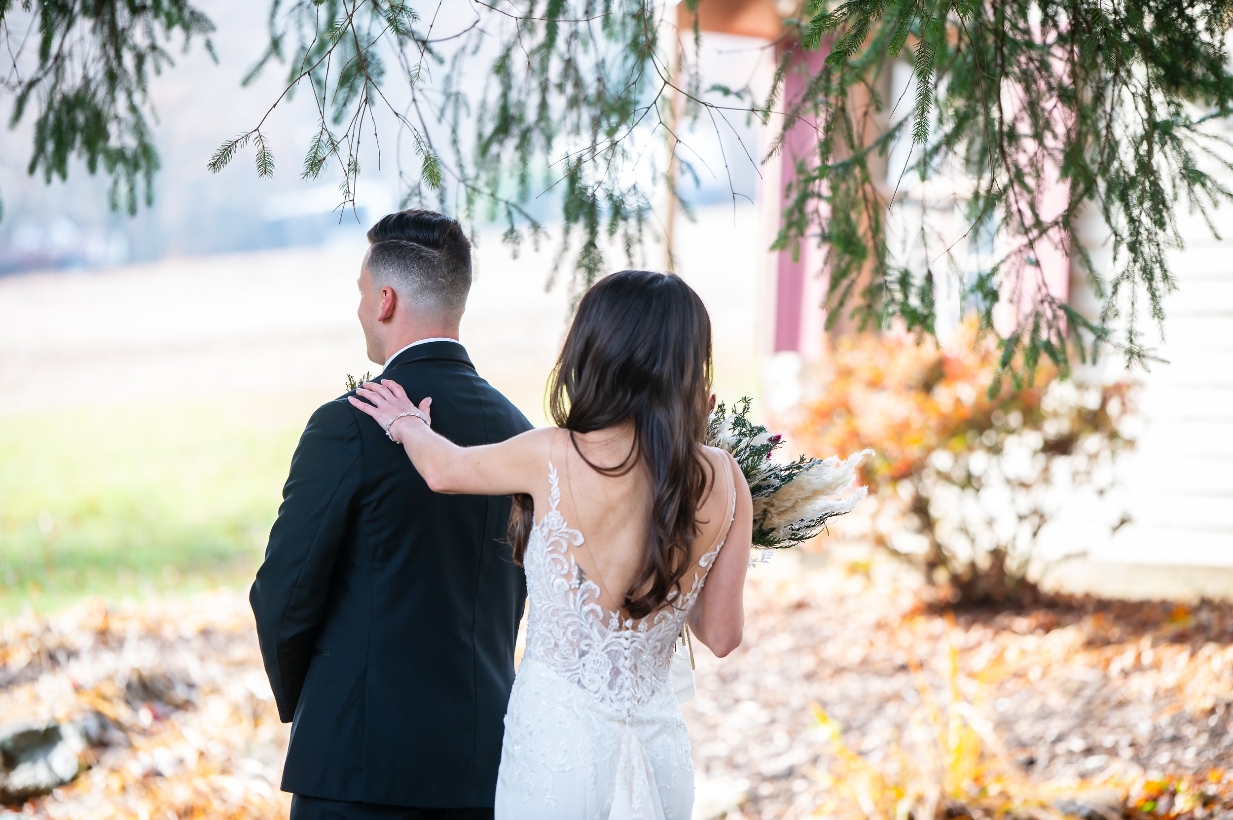 A bride and groom standing outdoors, with the bride resting her hand on the groom's shoulder, under a tree with autumn leaves in the background.