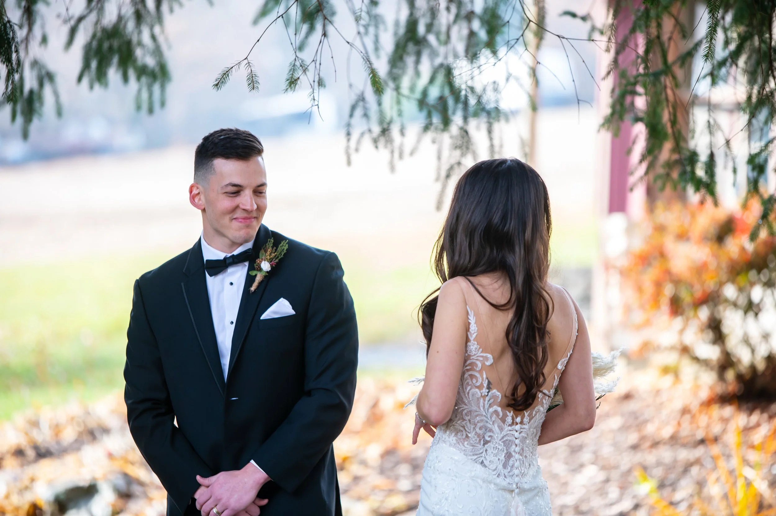 A groom in a black tuxedo with a boutonnière, smiling and looking at a bride in a white lace wedding dress with long dark hair, outdoors with trees and fall leaves in the background.