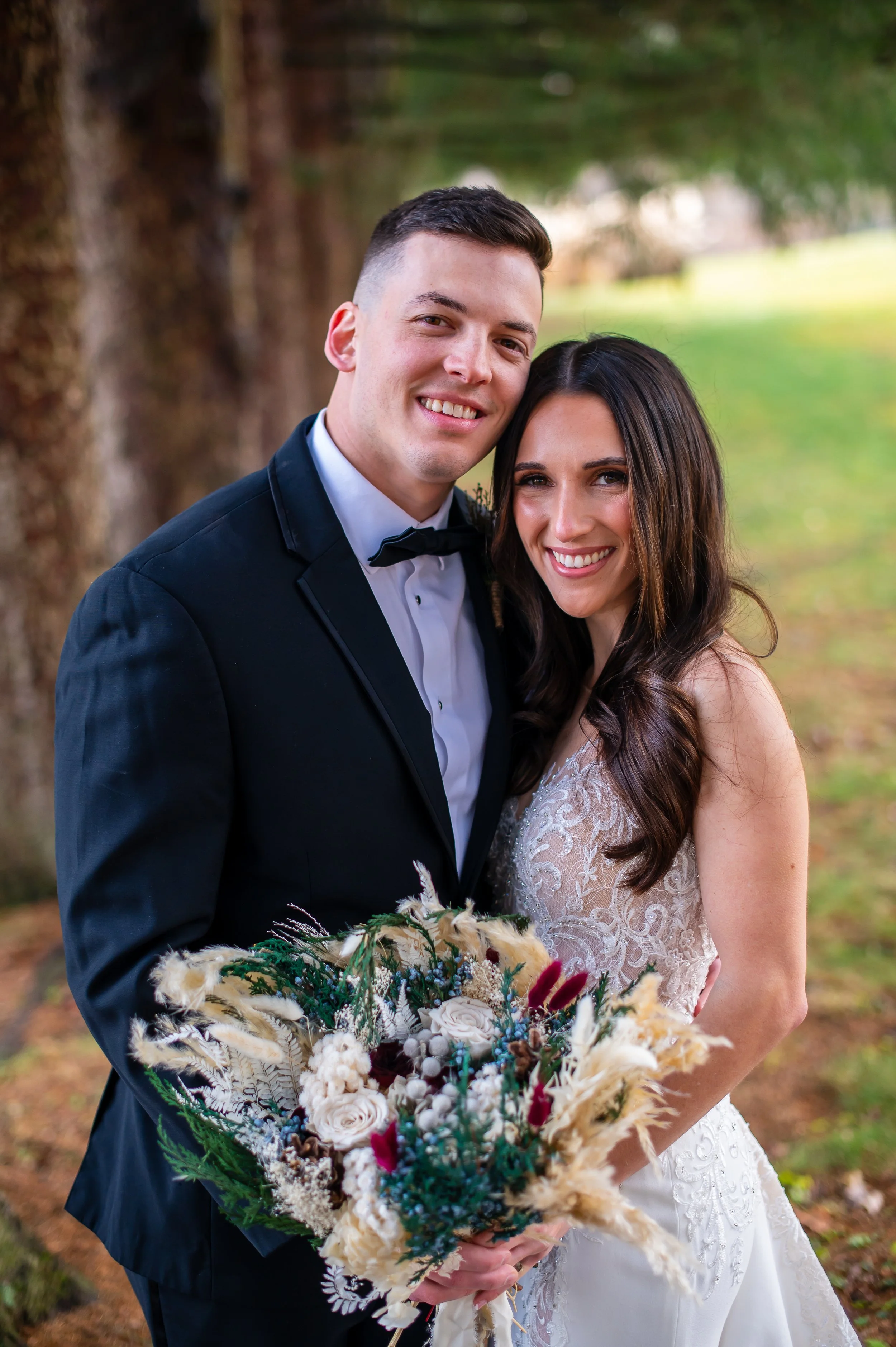 A newlywed couple stands outdoors on a grassy area, smiling, with the bride holding a large, colorful bridal bouquet. The groom is dressed in a black tuxedo with a bow tie, and the bride is wearing a lace wedding gown with long dark hair.