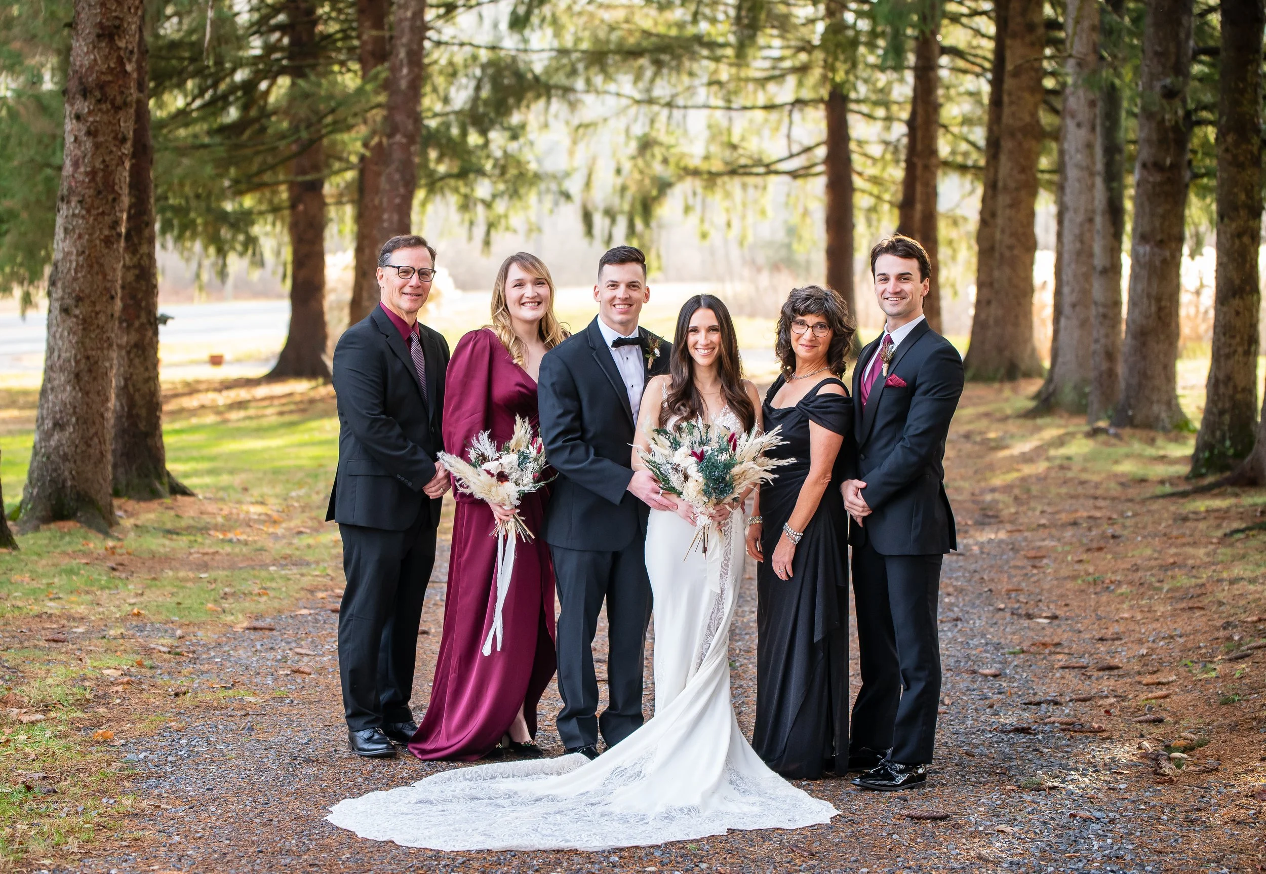A wedding group portrait outdoors featuring a bride in a white dress holding a bouquet, surrounded by five guests dressed in formal attire, standing on a forest path with tall trees and soft sunlight.