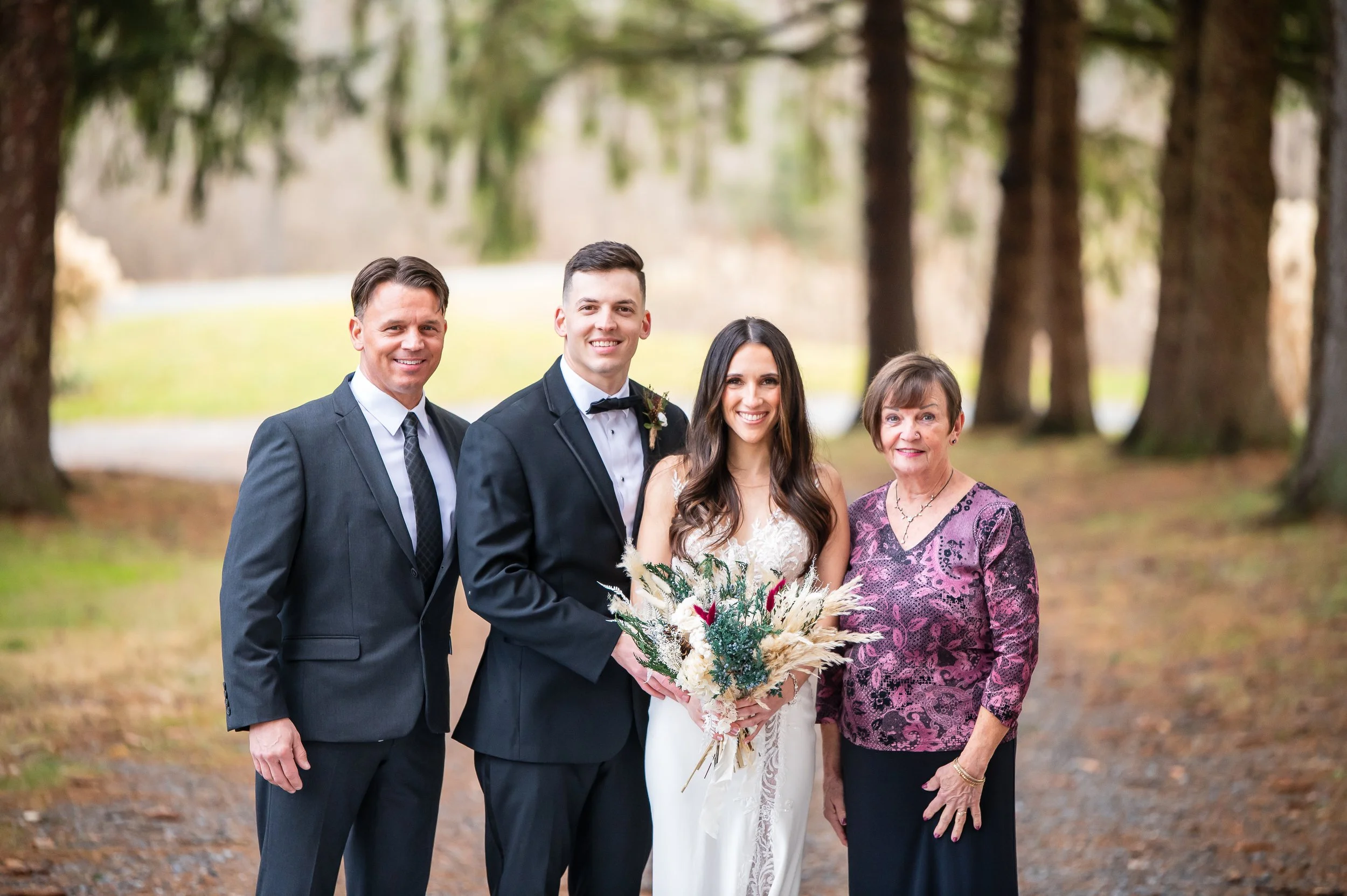 Group of four people, two men and two women, dressed in formal attire standing outdoors on a trail in a wooded area, smiling at the camera, with a woman in a white bridal gown holding a bouquet of flowers.