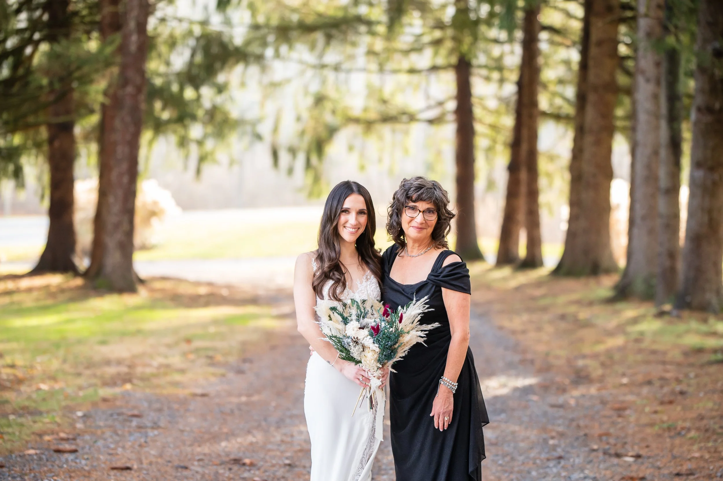 Two women in an outdoor setting with tall trees, one in a white wedding gown holding a bouquet, and the other in a black dress standing beside her.