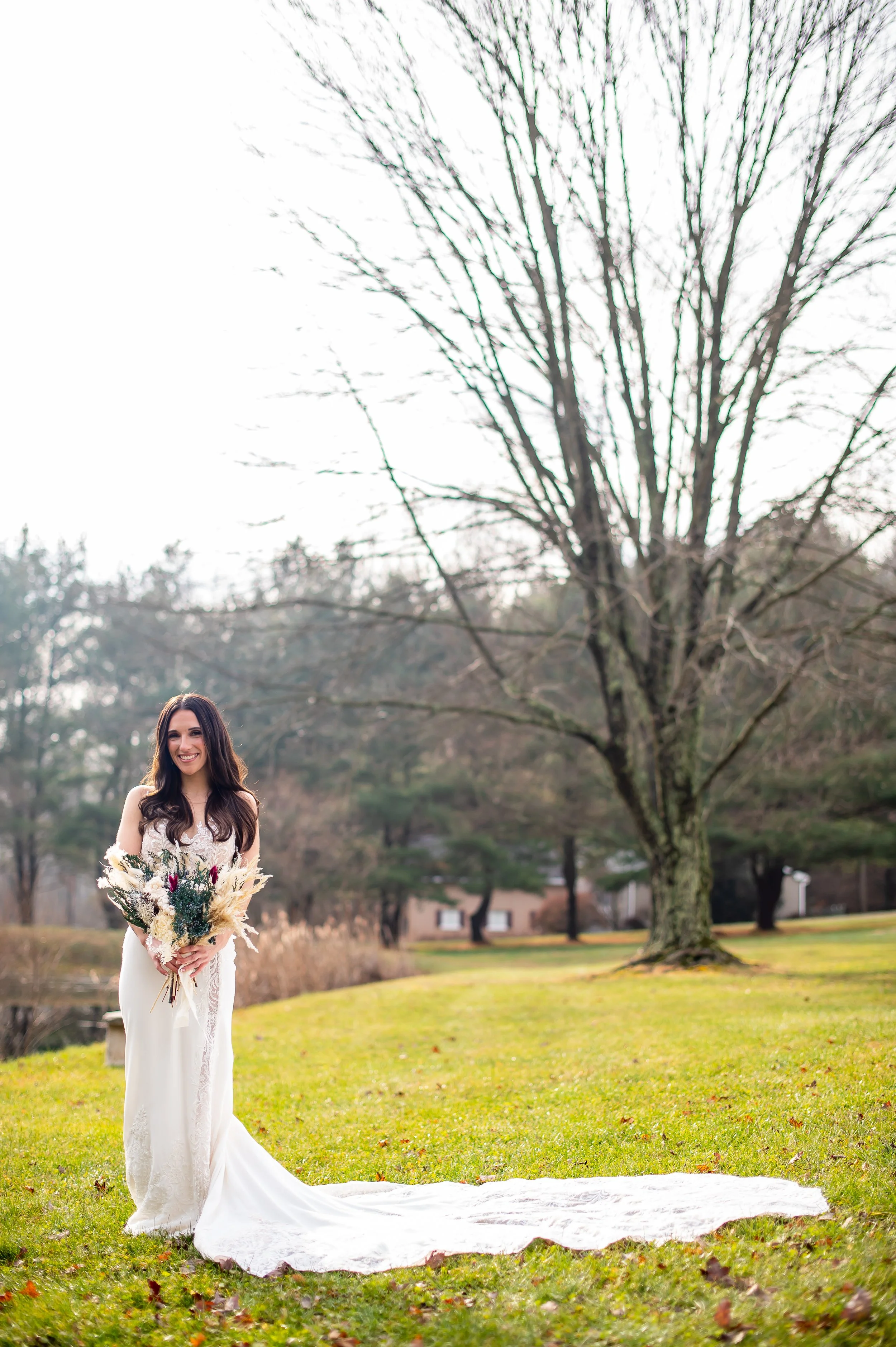 A woman in a white wedding dress holding a bouquet of flowers standing on a grass lawn with a large tree and houses in the background.