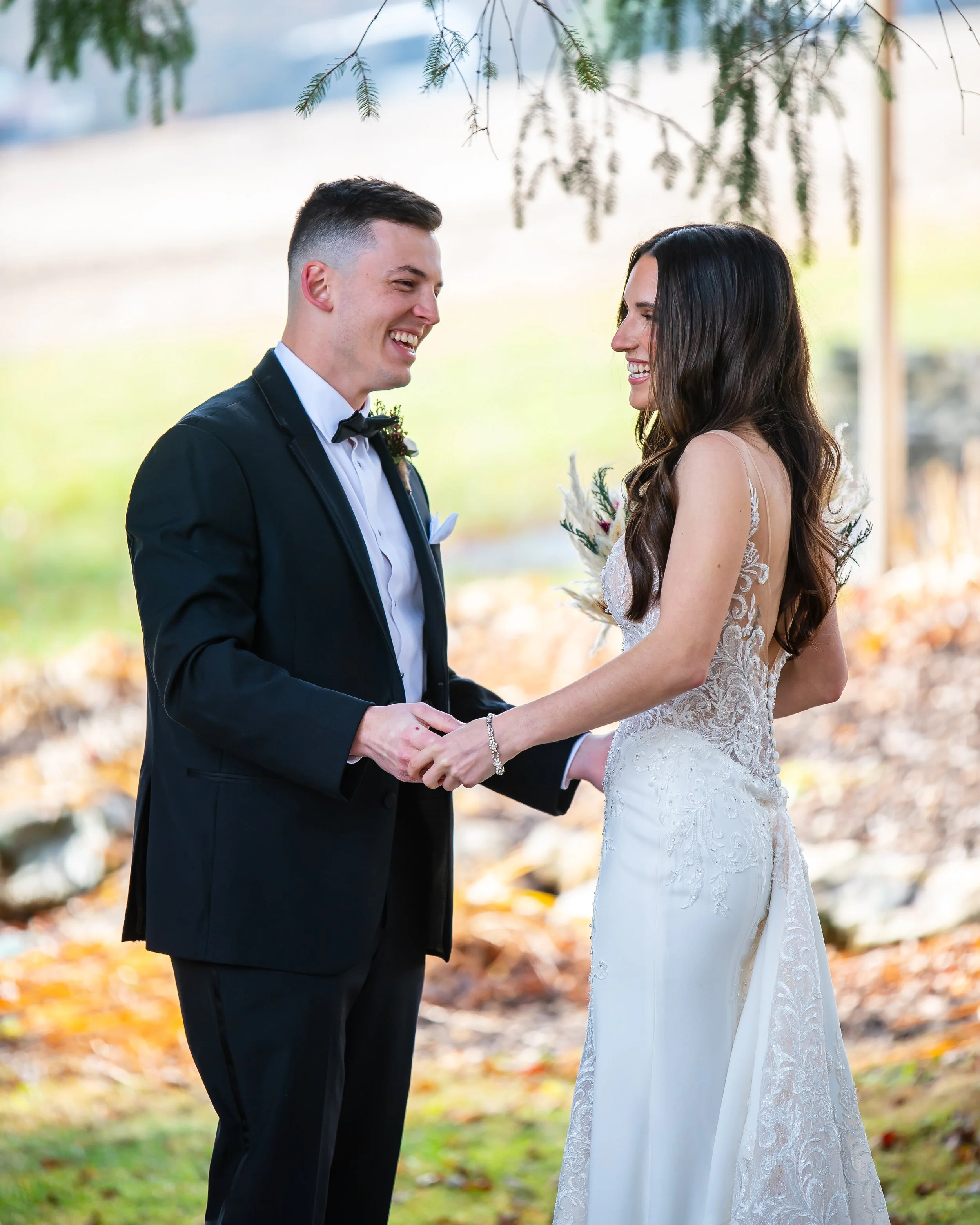 A couple in wedding attire exchanging vows outdoors with trees and fallen leaves in the background.