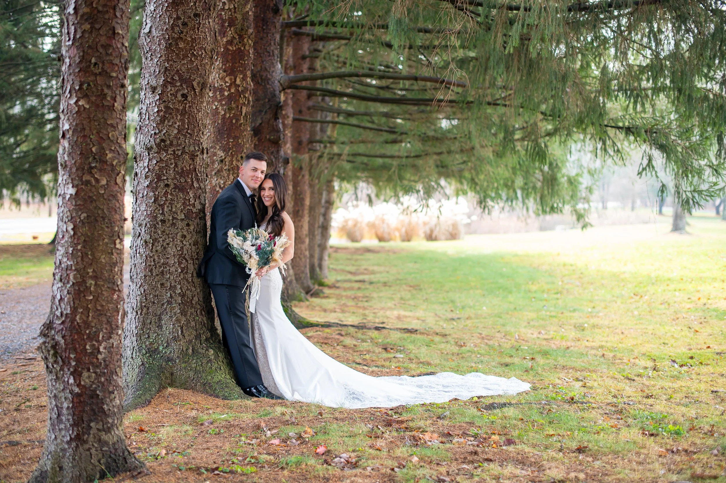 A newlywed couple dressed in wedding attire, standing close together and smiling, leaning against a large tree in a park with lush green grass and trees in the background.