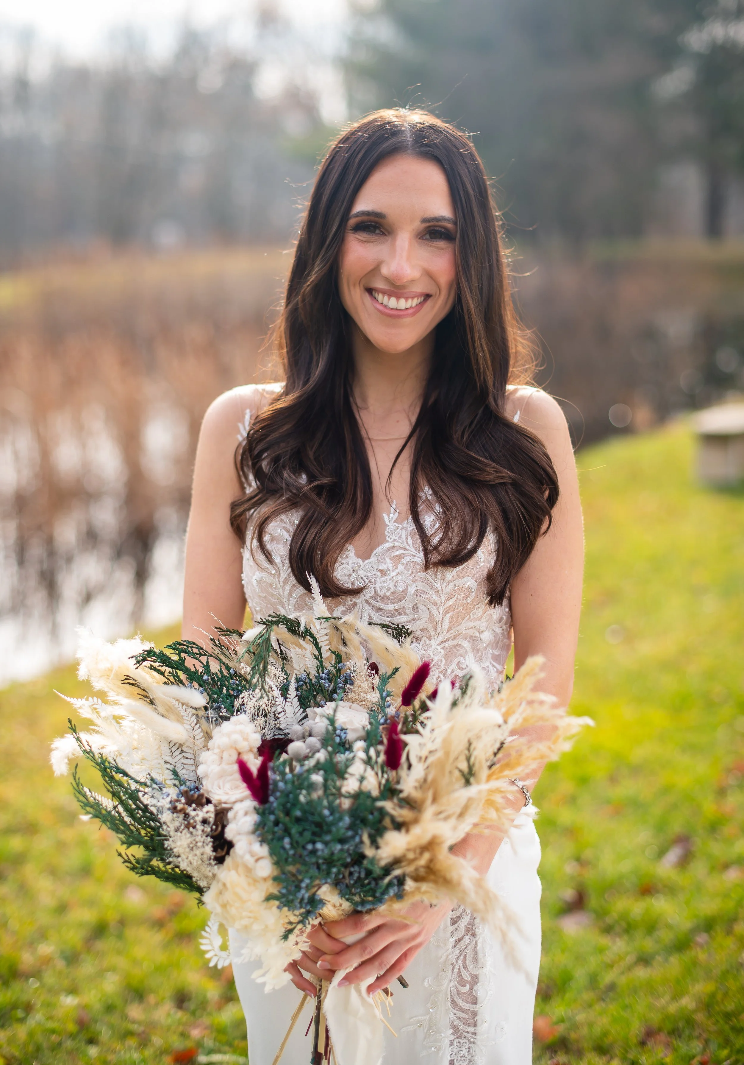 A woman in a wedding dress holding a bouquet of flowers outdoors near a pond, smiling at the camera.