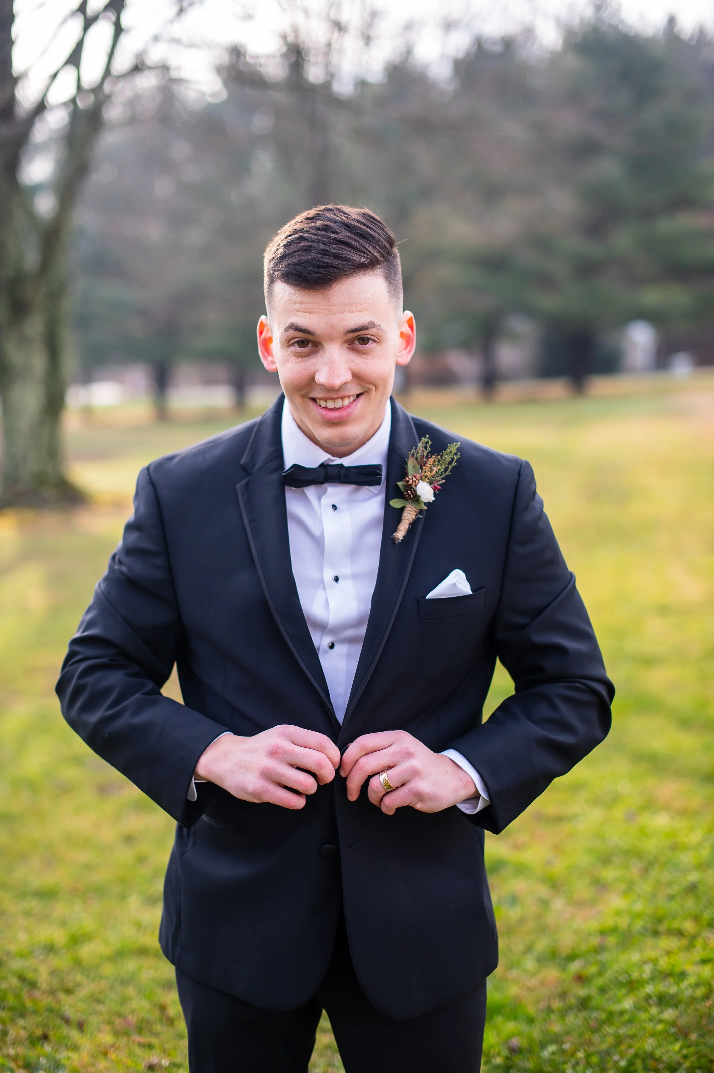 A young man in a black tuxedo with a bowtie, white dress shirt, and boutonniere on his lapel, standing outdoors on a grassy area with trees in the background. He is smiling and adjusting his jacket.