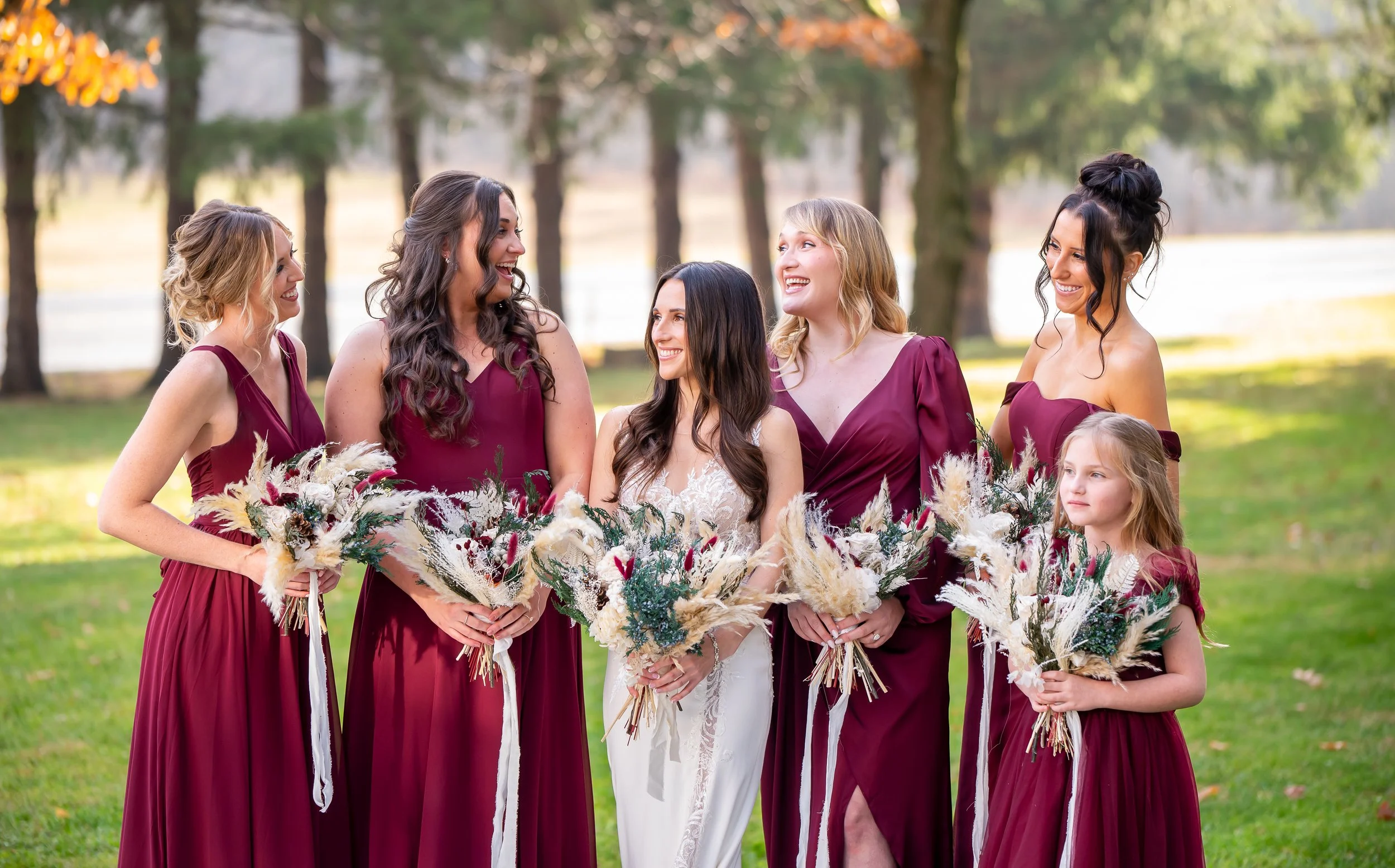 A bride and five bridesmaids standing outdoors on a grassy area with trees in the background, all holding bouquets and smiling at each other. The women are dressed in burgundy and white dresses.