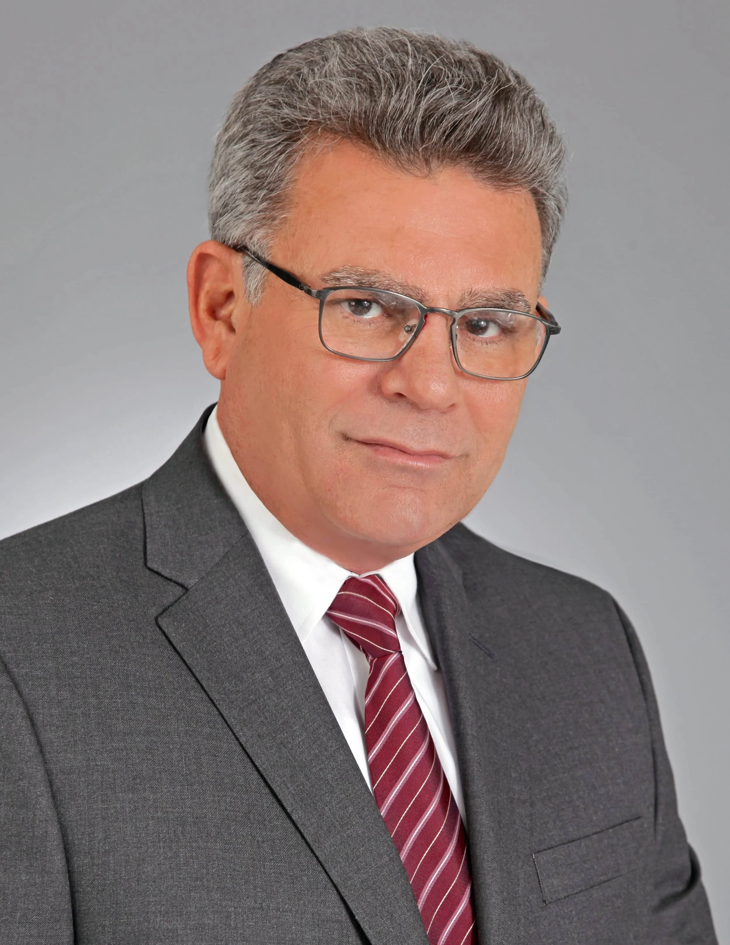 Man with gray hair and glasses wearing a gray suit, white shirt, and red striped tie against a plain gray background.