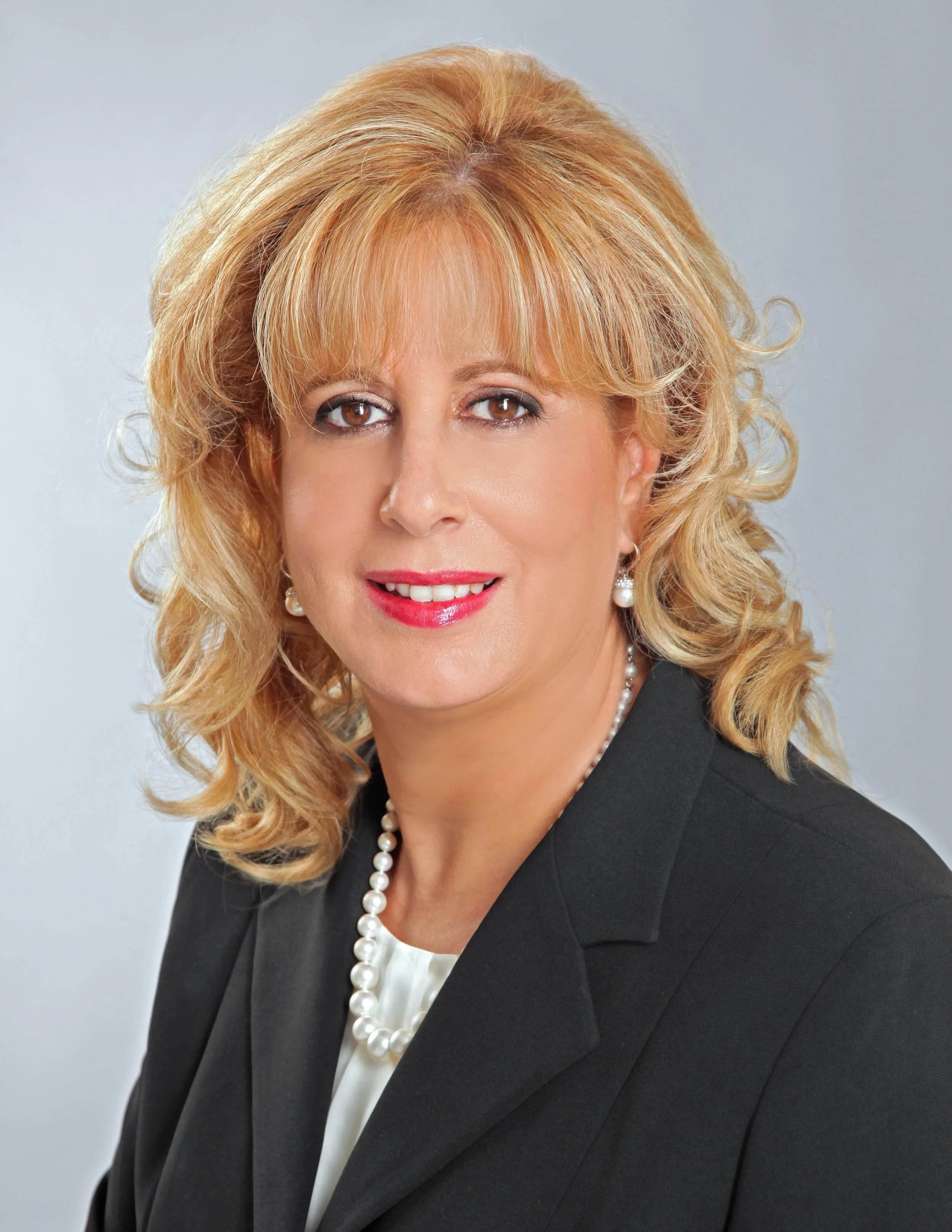 A woman with blonde curly hair in a black blazer, white top, pearl necklace, and earrings, smiling at the camera.