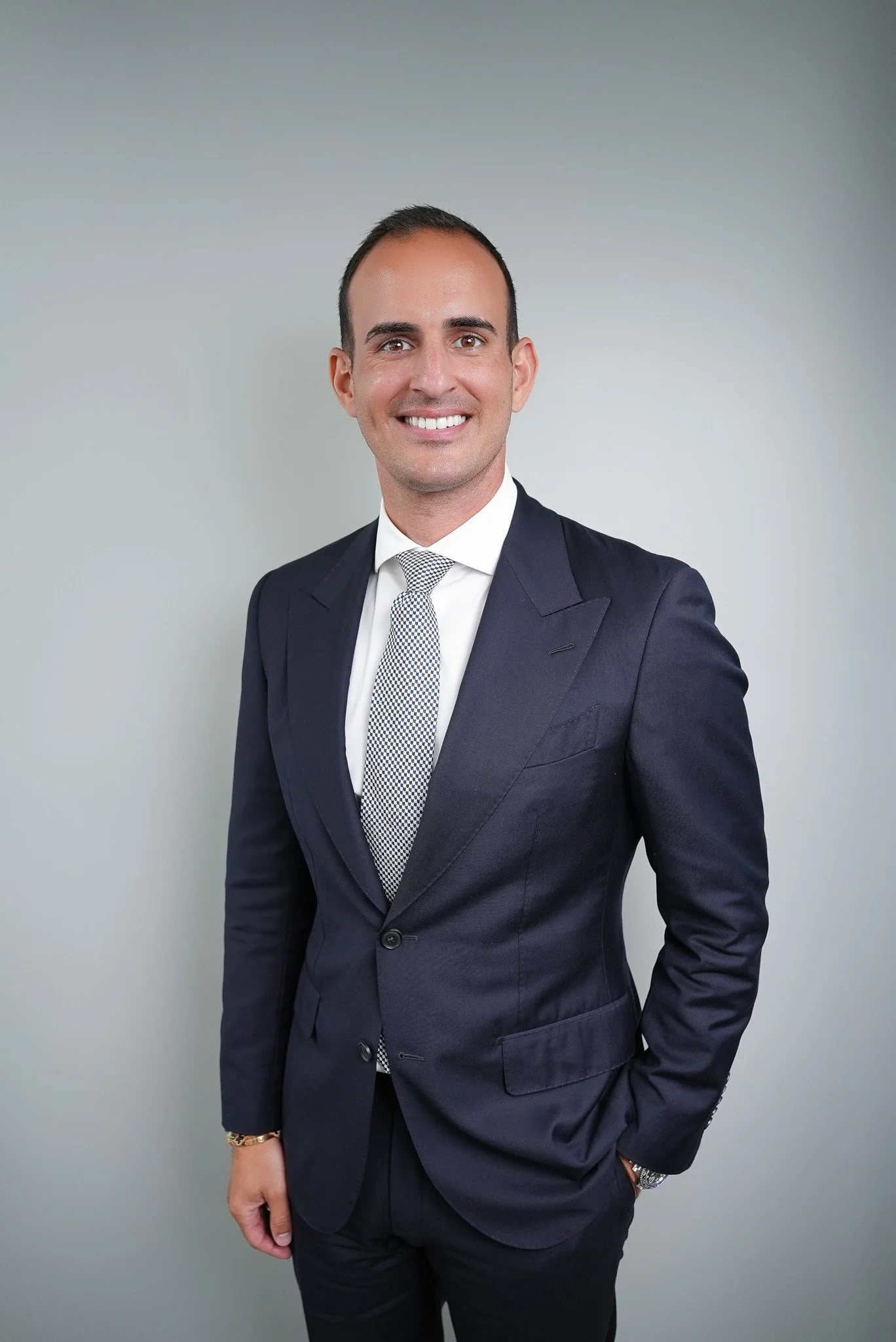 A man in a dark suit and tie stands smiling against a plain light gray background.