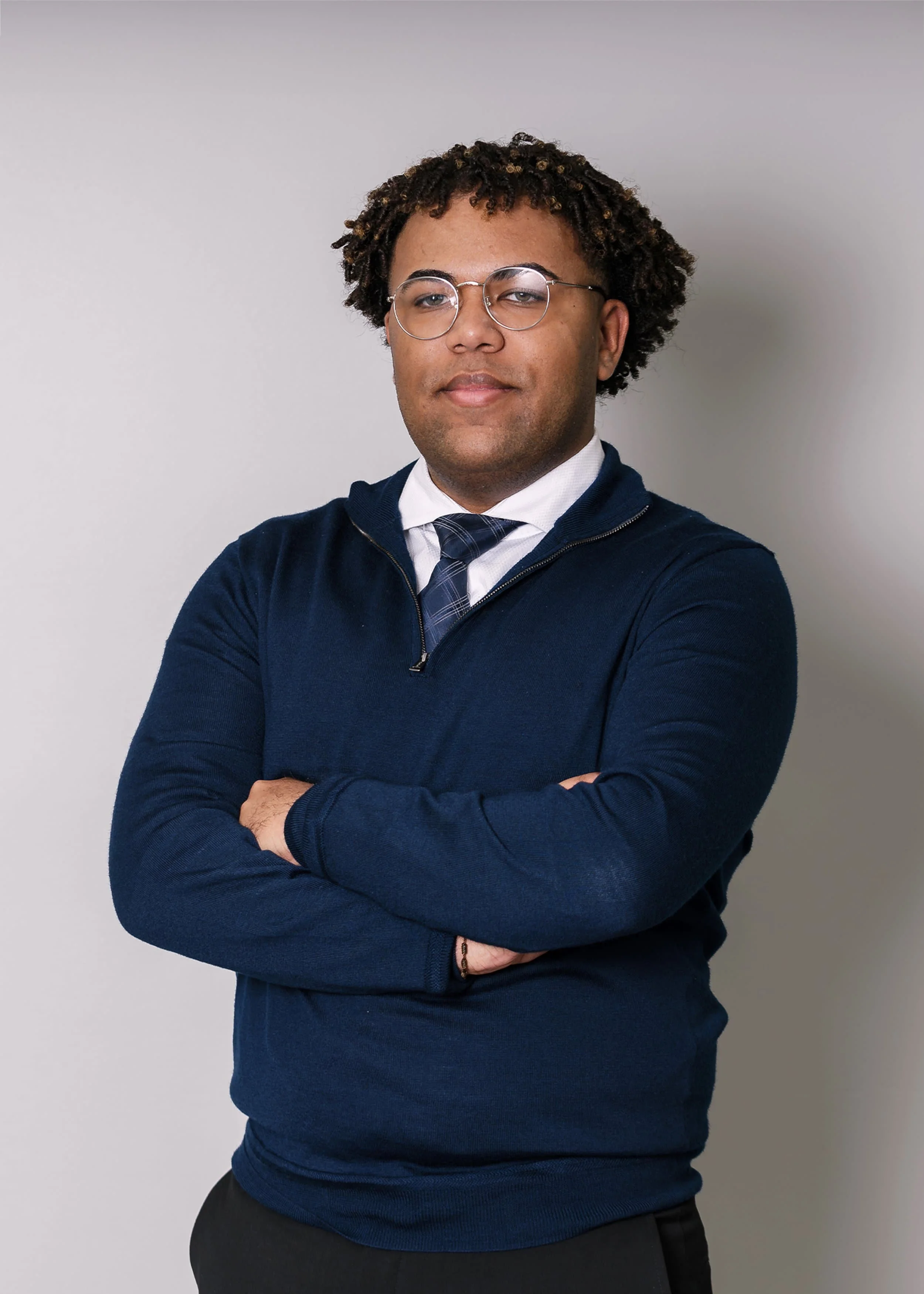 A person with glasses and curly hair, wearing a blue sweater and tie, stands with arms crossed against a plain background.
