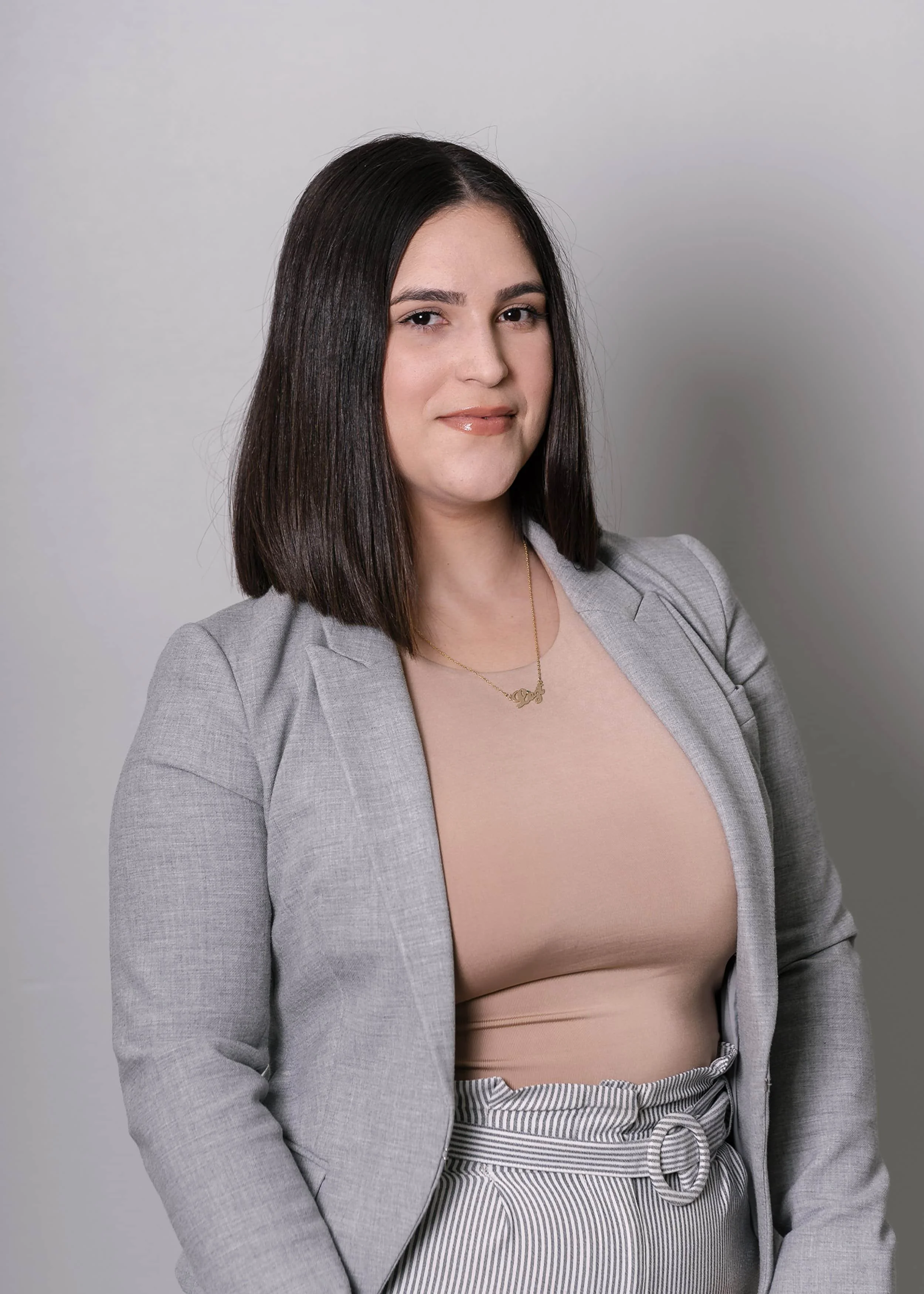 Woman with straight dark hair in a gray blazer, beige top, and striped pants, standing against a plain light background, smiling.