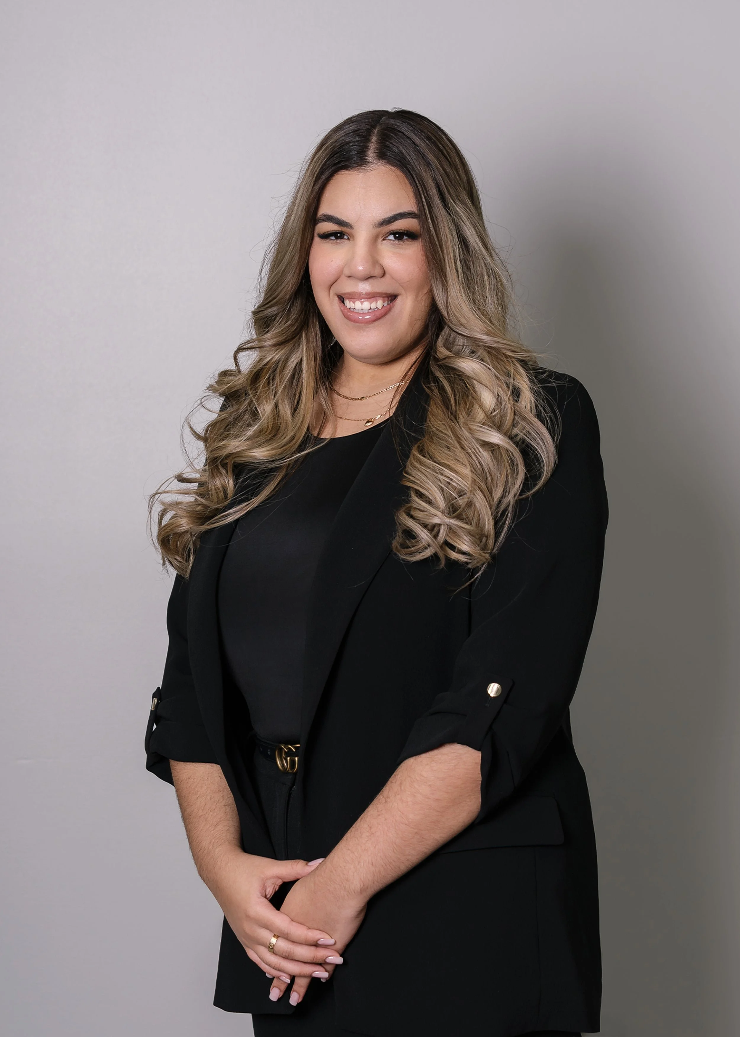 Smiling woman with long wavy hair in a black blazer stands against a plain light gray background.