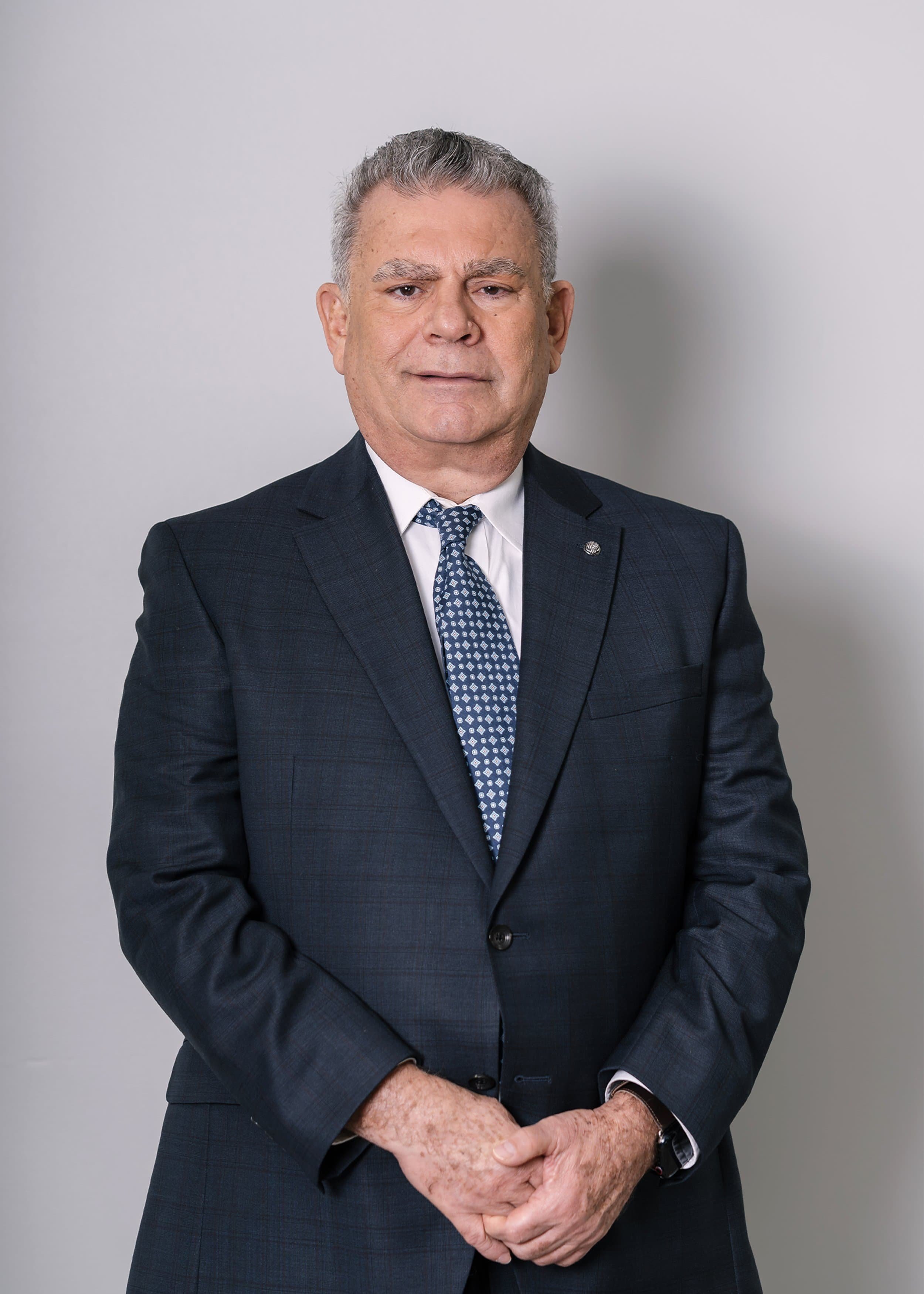 Man in a dark suit and patterned tie stands against a plain light background, hands clasped in front, looking at the camera.