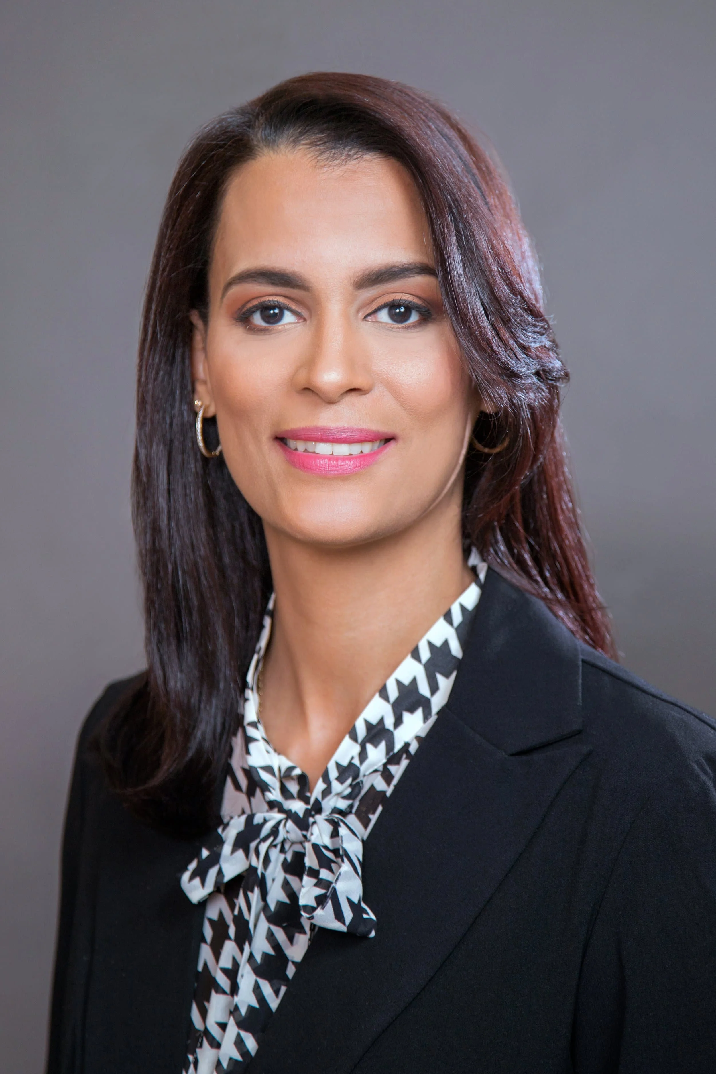 Woman with straight dark hair in a blazer and patterned blouse, smiling against a plain gray background.