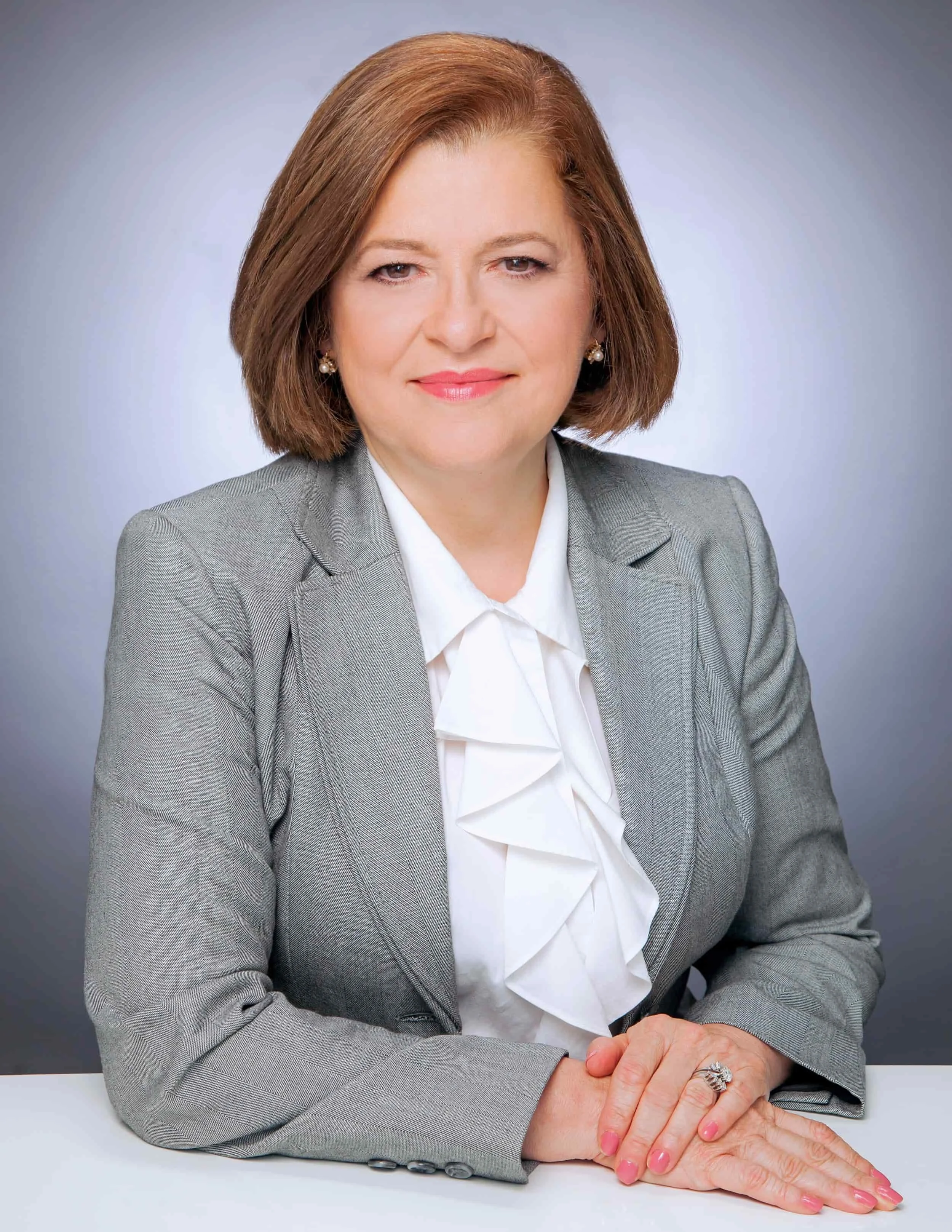 Woman with short brown hair in a gray blazer and white blouse, sitting at a table, smiling with hands folded.