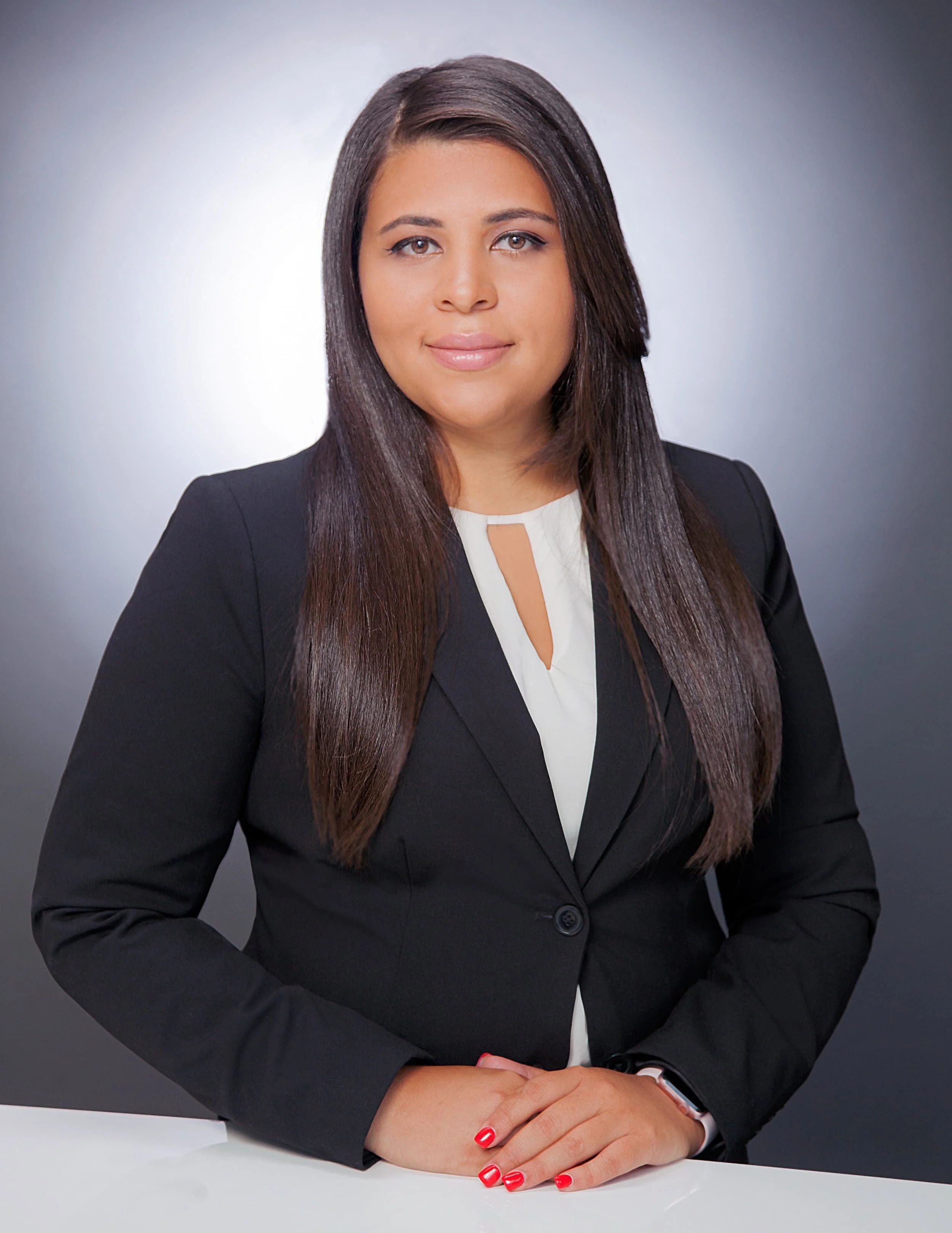 Woman in a black blazer and white blouse with long straight hair, sitting at a desk and smiling in a professional portrait.