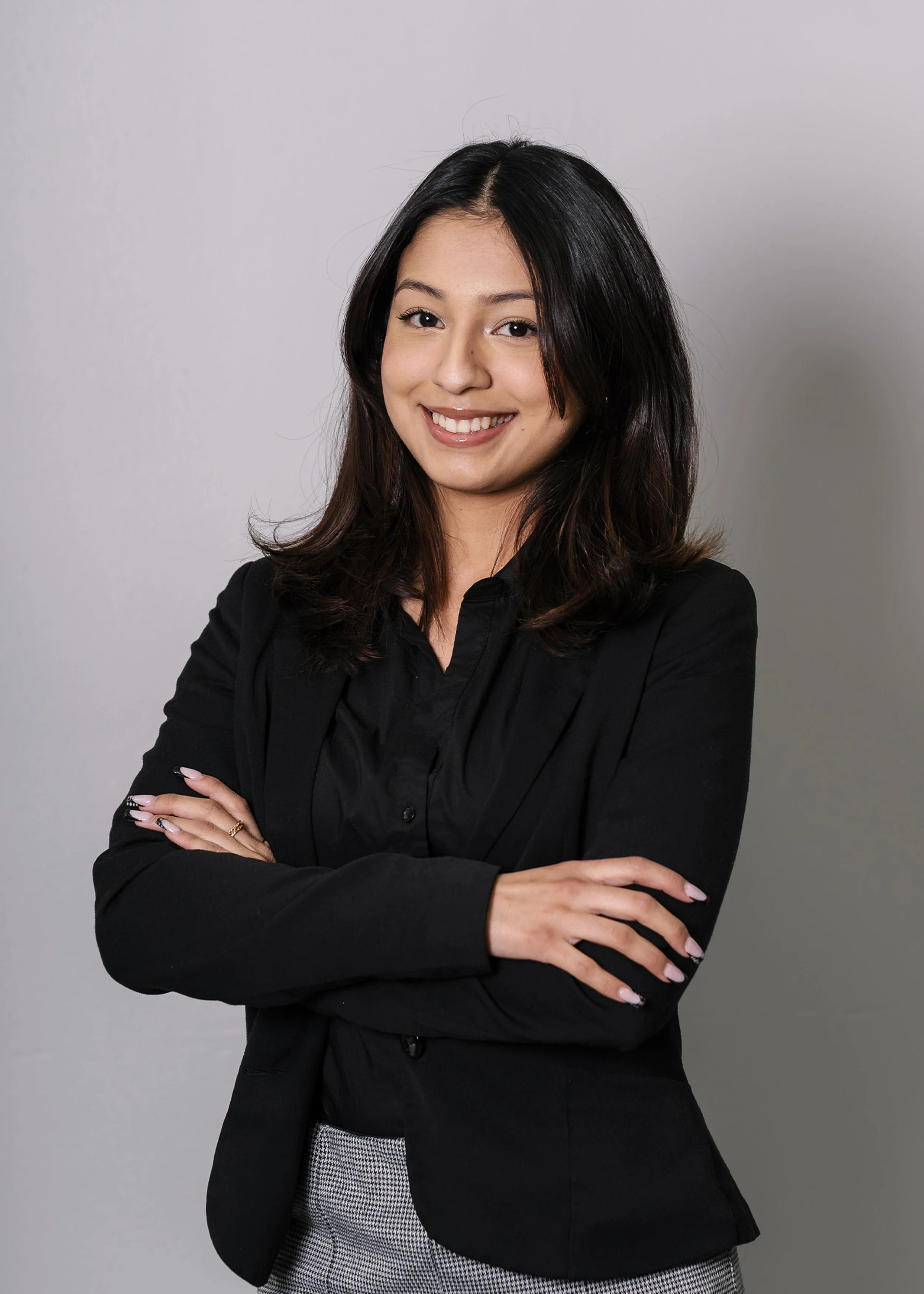 A woman with long dark hair, wearing a black blazer and checked pants, smiles with arms crossed against a plain background.