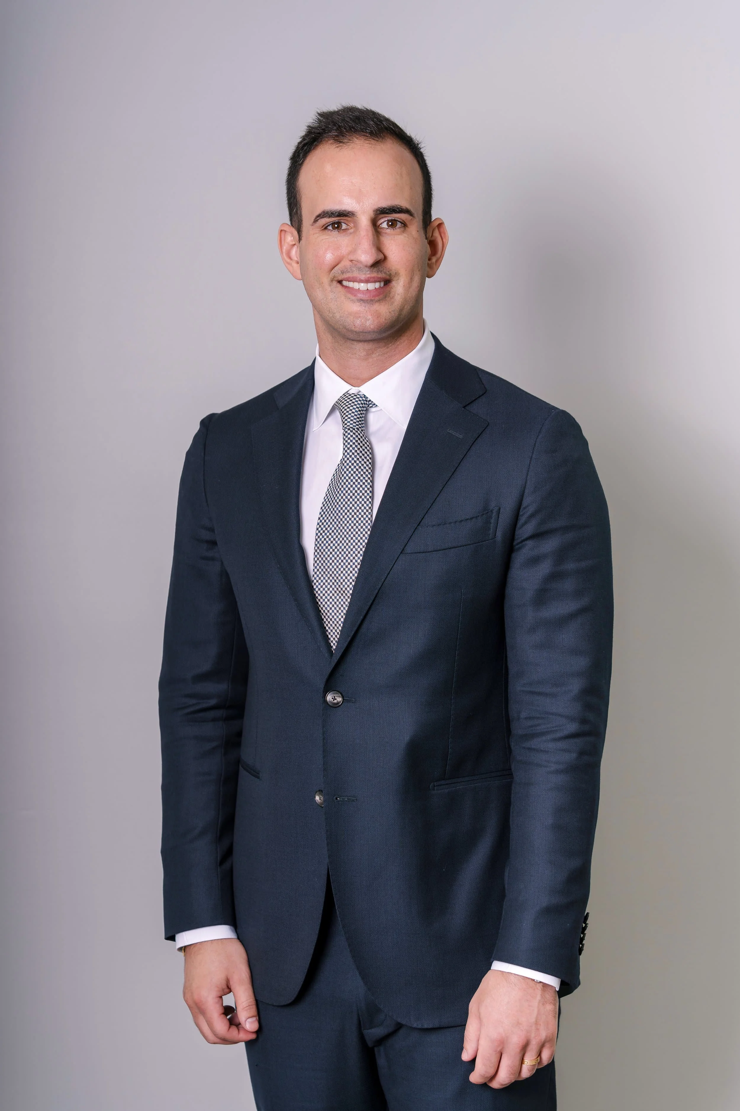 A man in a dark suit and tie stands smiling against a plain light gray background.