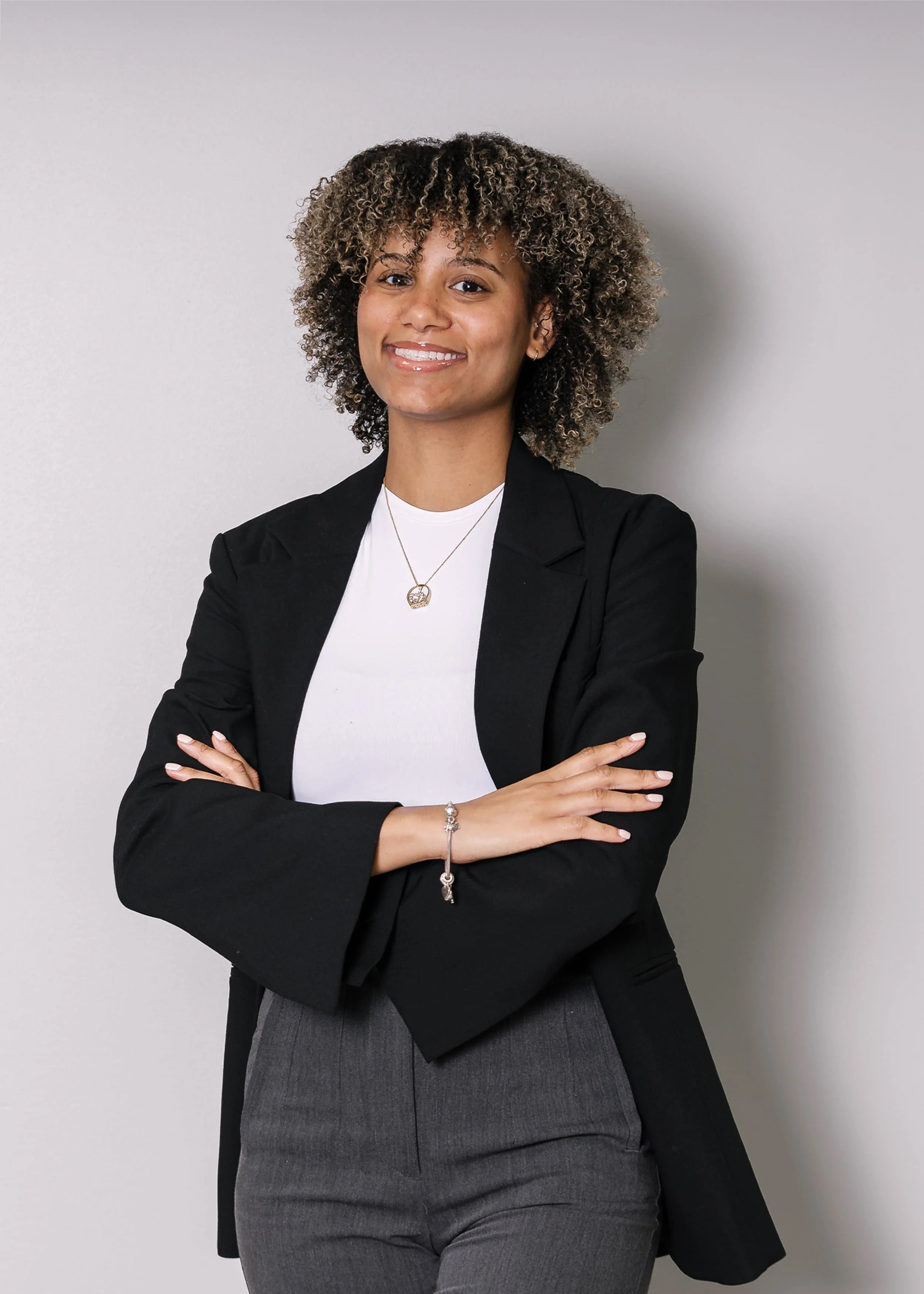 A woman with long dark hair in a black blazer and white shirt smiles in front of a plain, light gray background.
