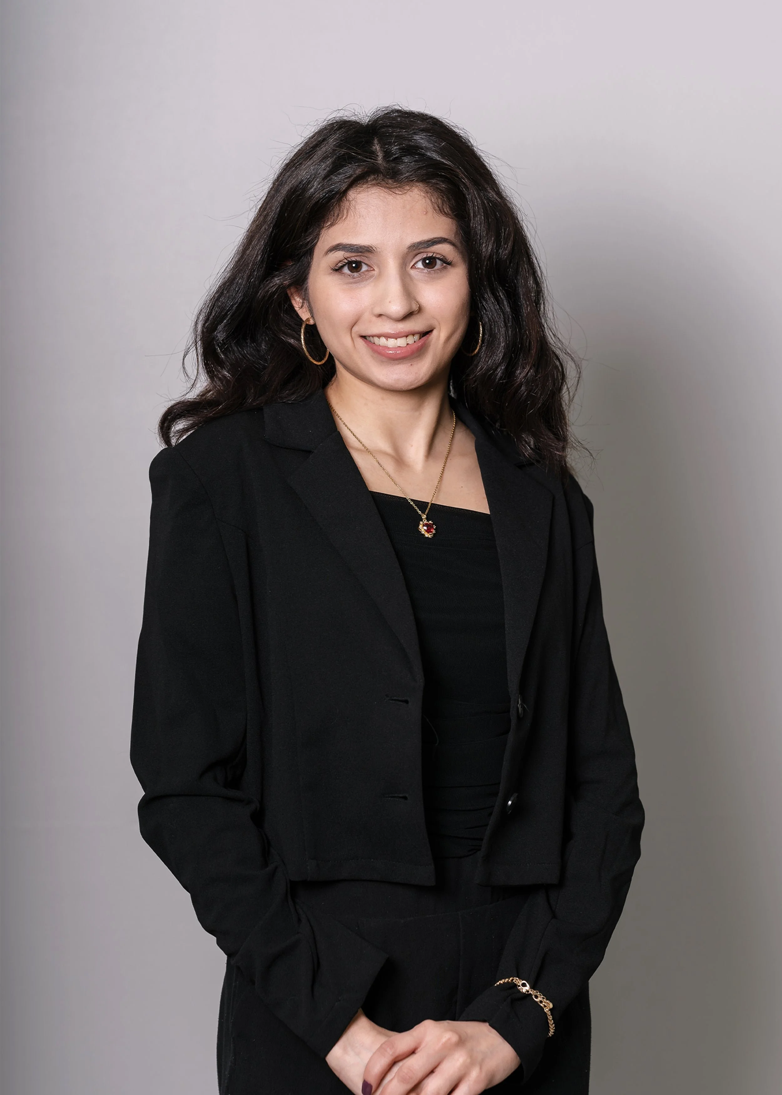 Young woman with long dark hair in a black suit, standing and smiling in front of a plain light gray background.