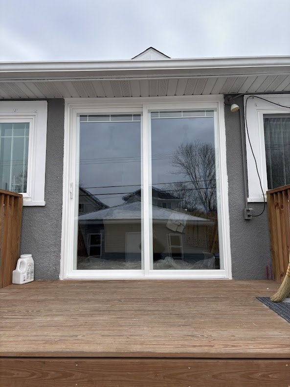 Photo of a backyard deck with sliding glass patio door leading inside a house, with a gray exterior wall and a small portion of a wooden fence visible.