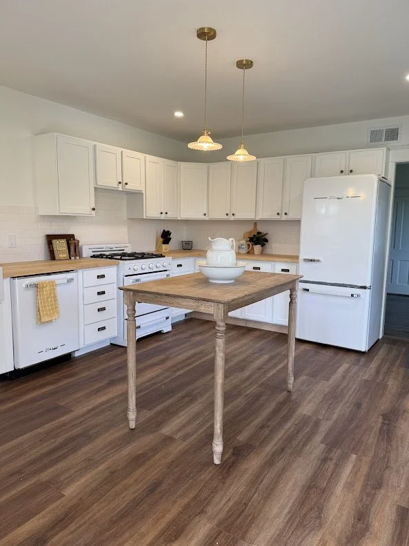 Kitchen with white cabinets, a wooden kitchen table with a white pitcher and bowl on top, hardwood floors, and two hanging pendant lights.