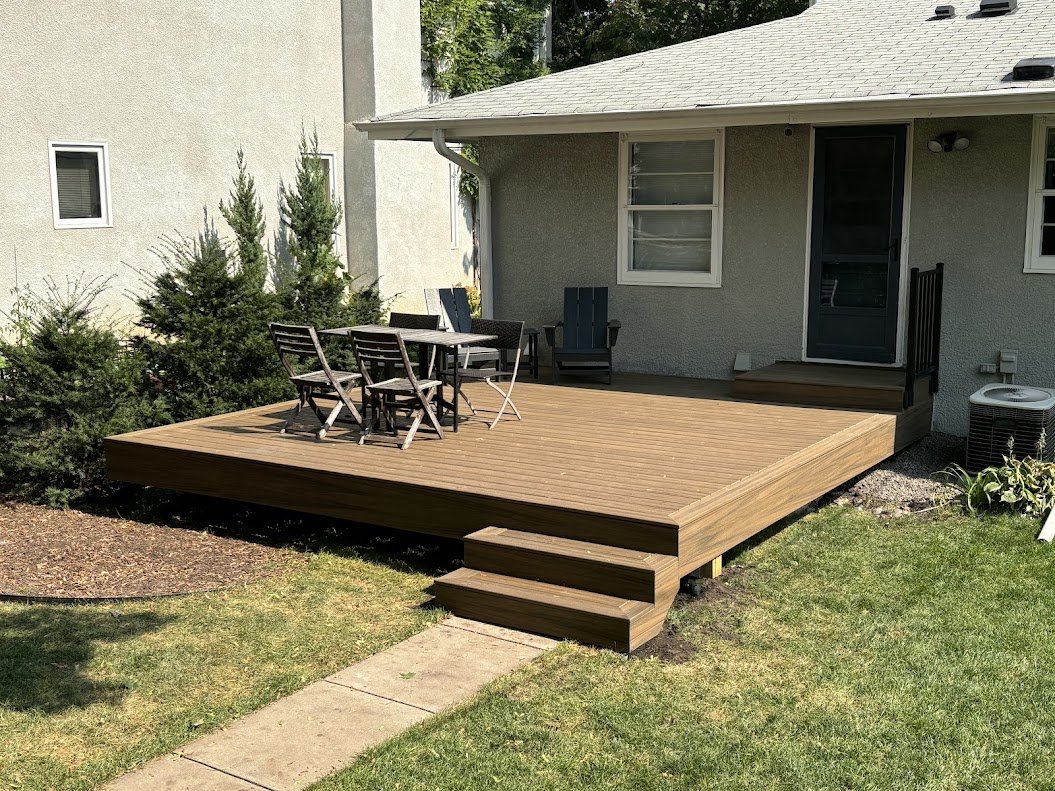 Wooden deck with outdoor furniture consisting of six chairs and a table, attached to a gray house with a door and windows, surrounded by grass and bushes.