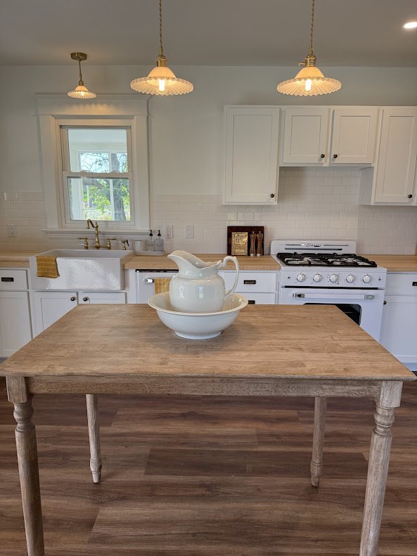 Kitchen with white cabinets, a window above the sink, and a wooden table in the center holding a pitcher and bowl. There are three hanging pendant lights and a gas stove against the wall.