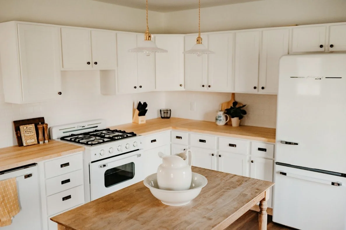 White kitchen cabinets with wooden countertops, white appliances including a refrigerator and stove, a kitchen island with a pitcher and bowl, hanging pendant lights, and decorative items like plants and jars.