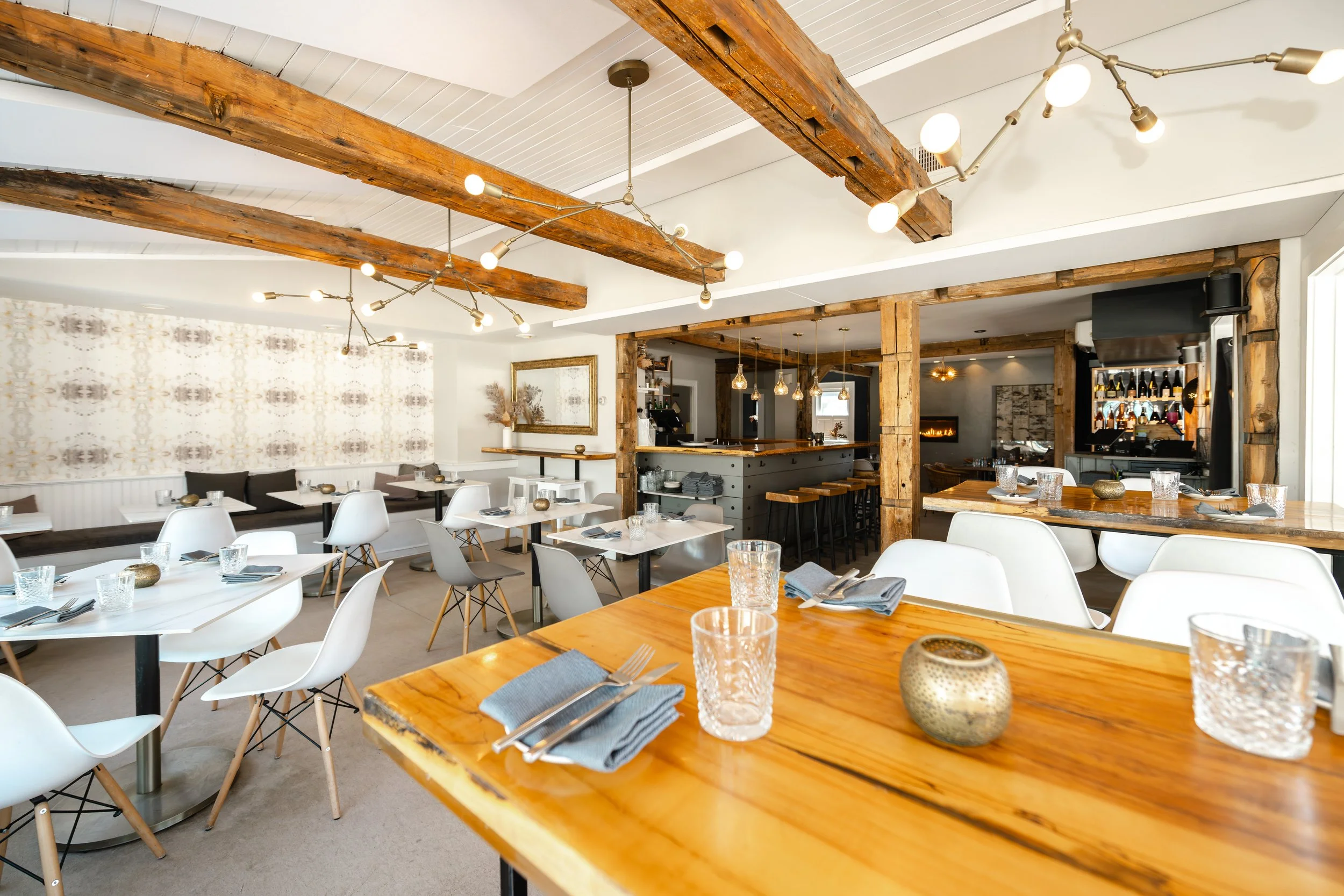 Interior of a modern restaurant with white tables, white chairs, wooden beams on the ceiling, warm lighting, and a bar area in the background.