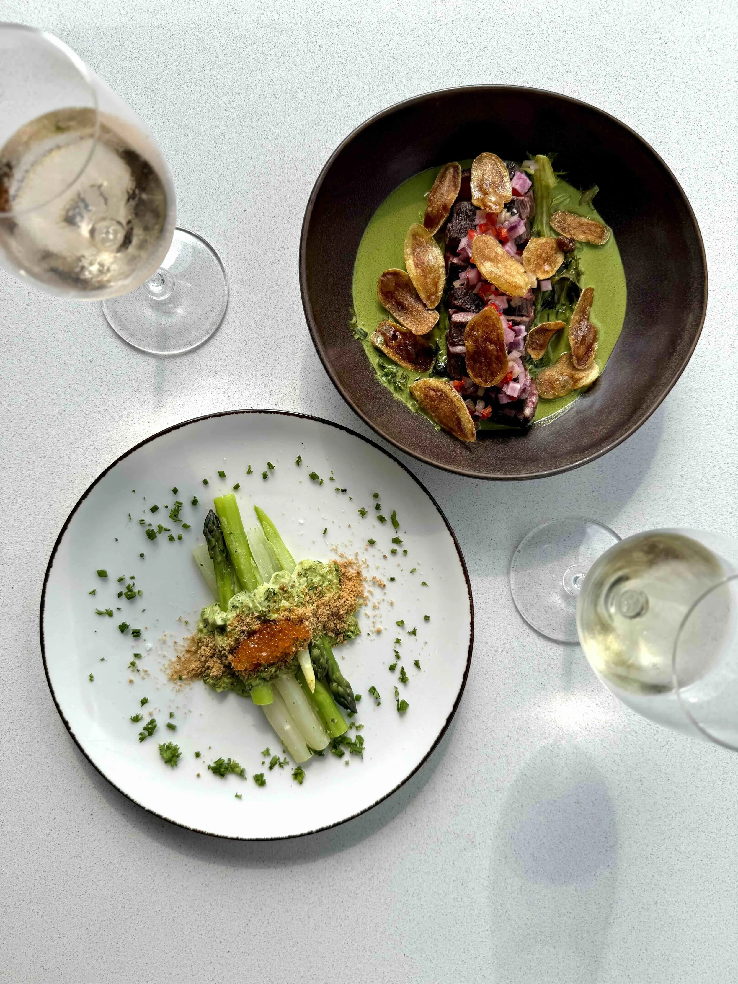 A plate of local beef, greens, and chips as a large plate. A small plate featuring asparagus, breadcrumbs, and fish row. Two glasses of Wine.