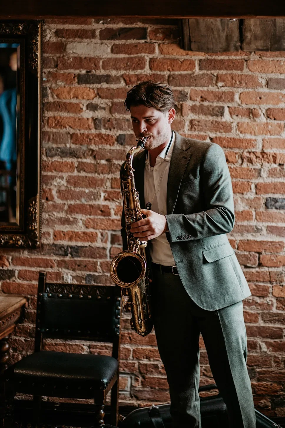 A young man in a gray suit playing a saxophone indoors against a brick wall.